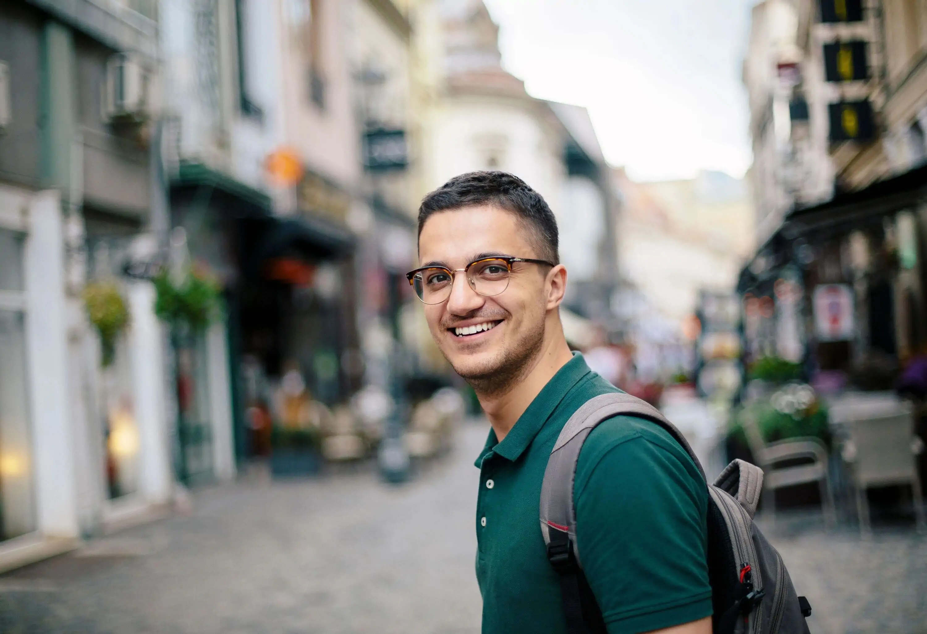 A man in eyeglasses and dark green shirt smiles as he crosses a street.