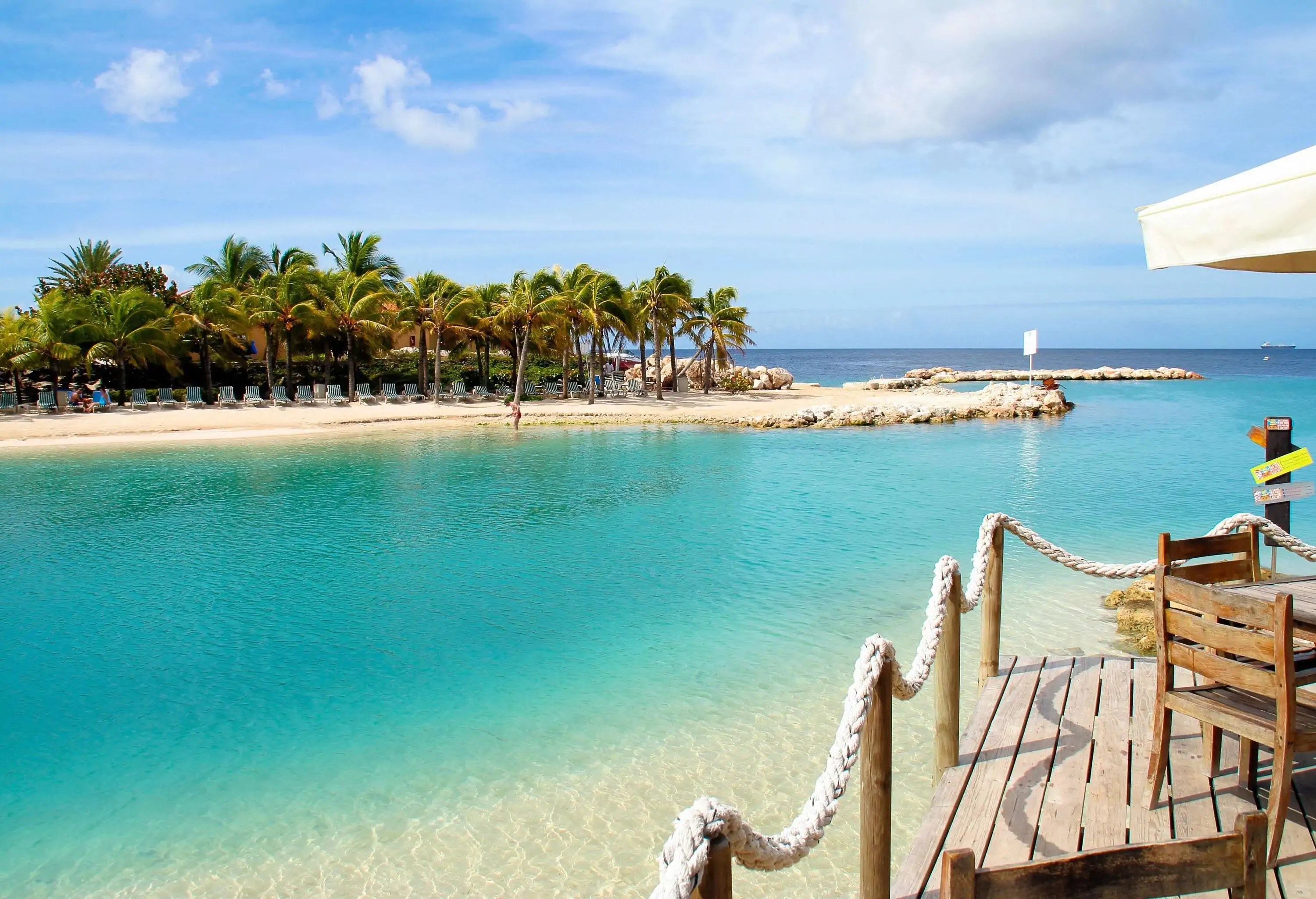 A luxurious white sand shore lined with sun loungers backed by tall tropical trees, all surrounded by the turquoise sea under the cloudy blue sky.