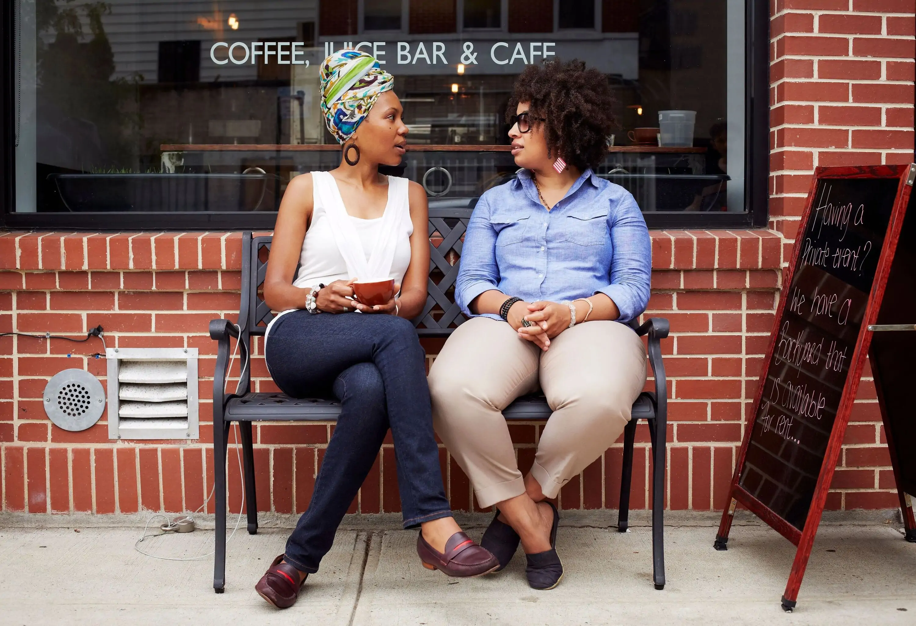 Two people talking on a bench outside of a coffee shop.
