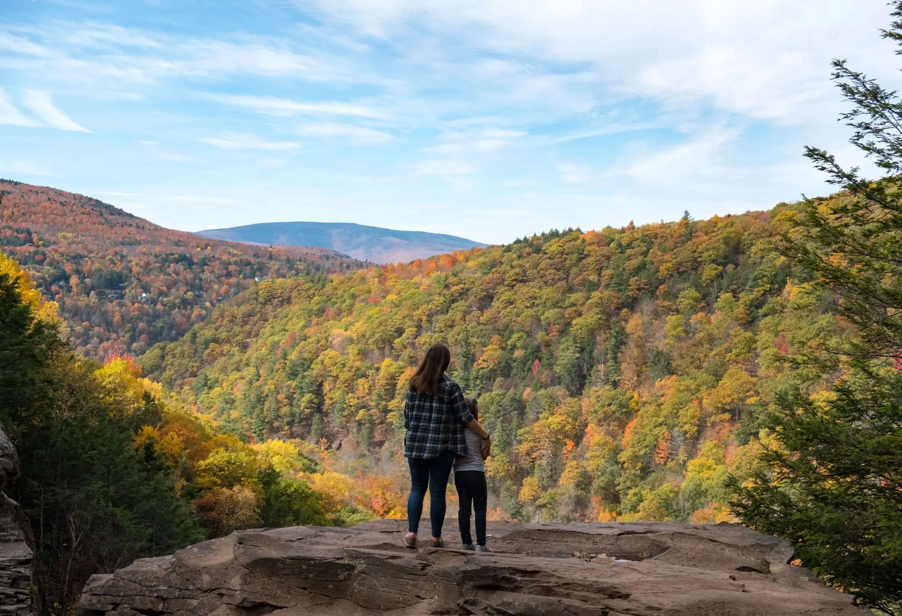 A mother and child embrace while standing on a rock ledge and admiring the mountains' vibrant autumnal foliage.