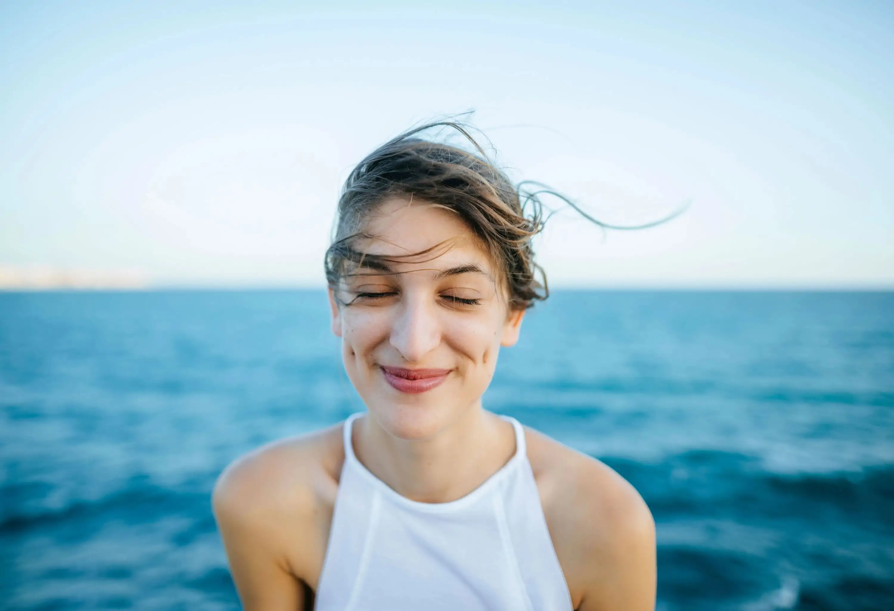 A smiling woman with closed eyes with the sea in the background.
