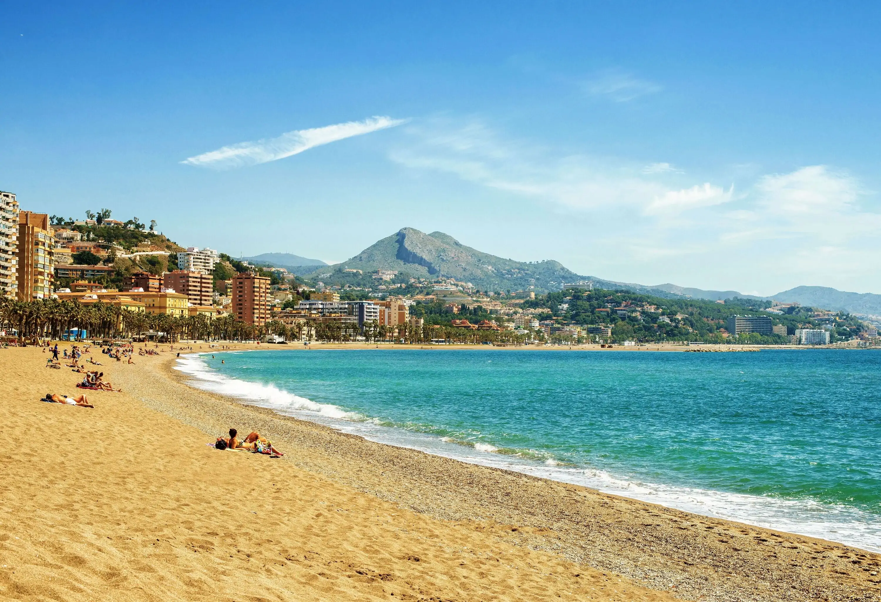 A tranquil blue beach with a sandy shore lined with modern buildings with a view of a mountain against the clear blue sky.