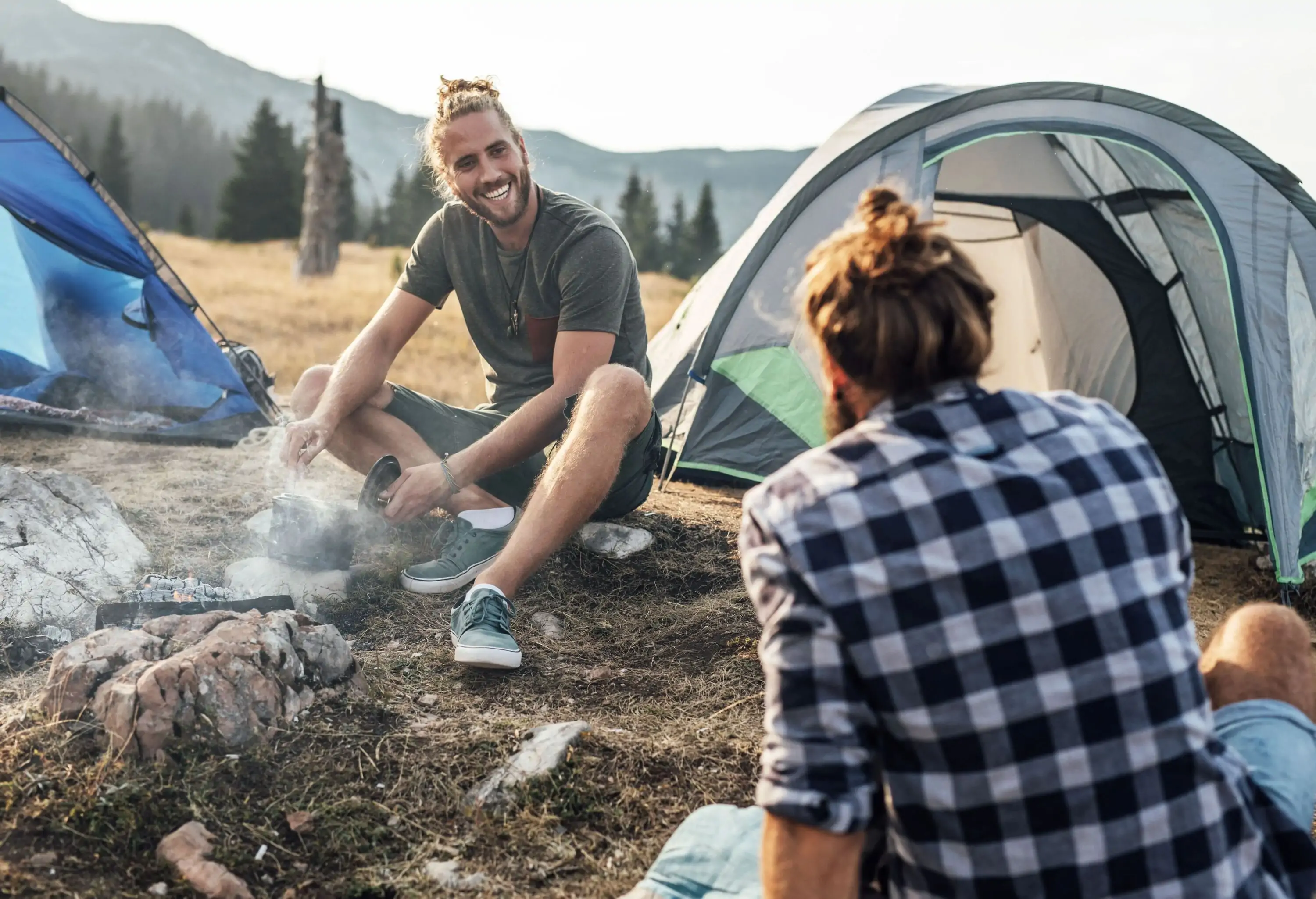 Two men are sitting on the ground by a campfire next to their camping tents.