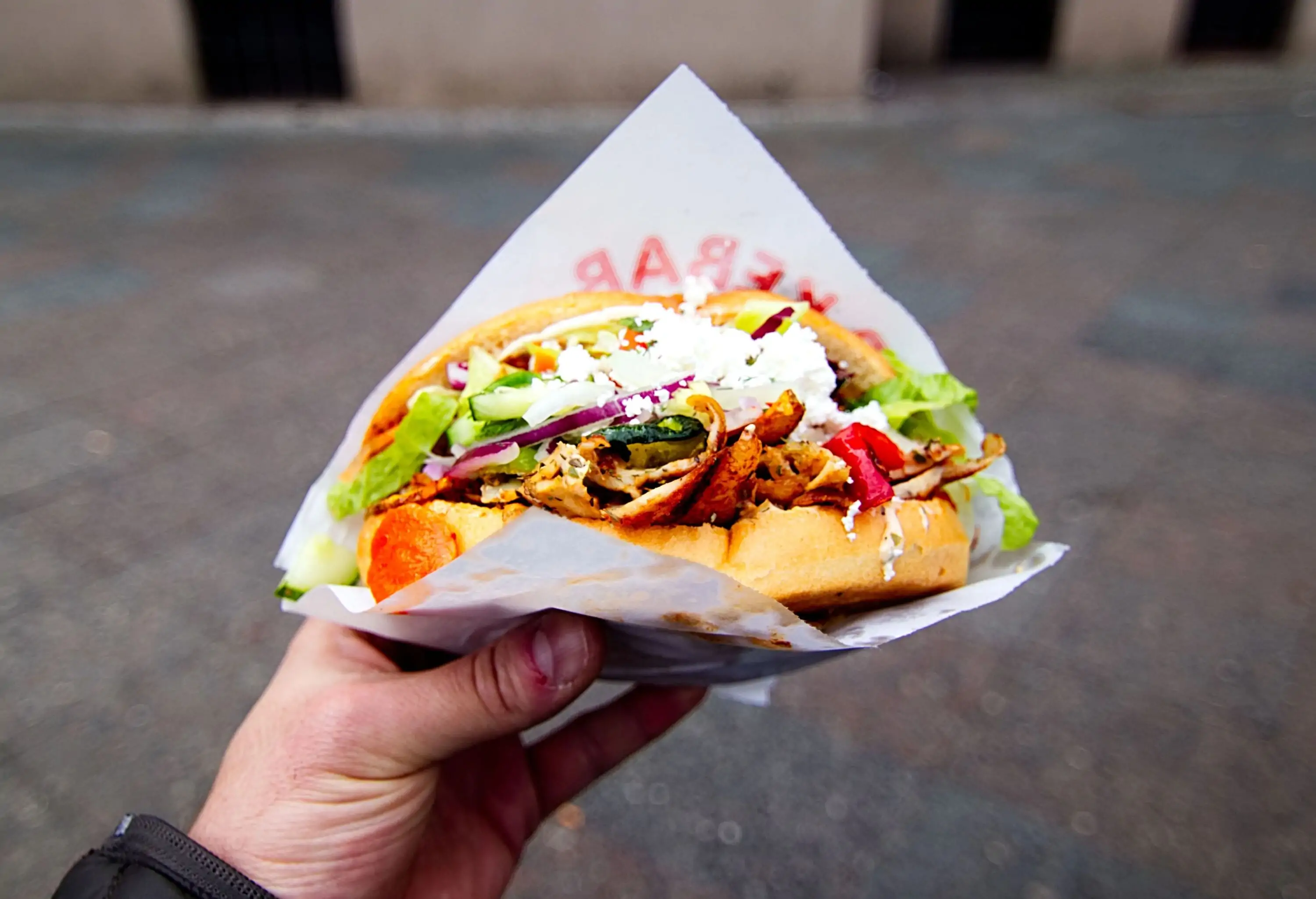 A hand holds a sandwich with meat, vegetables, and cheese wrapped in paper.