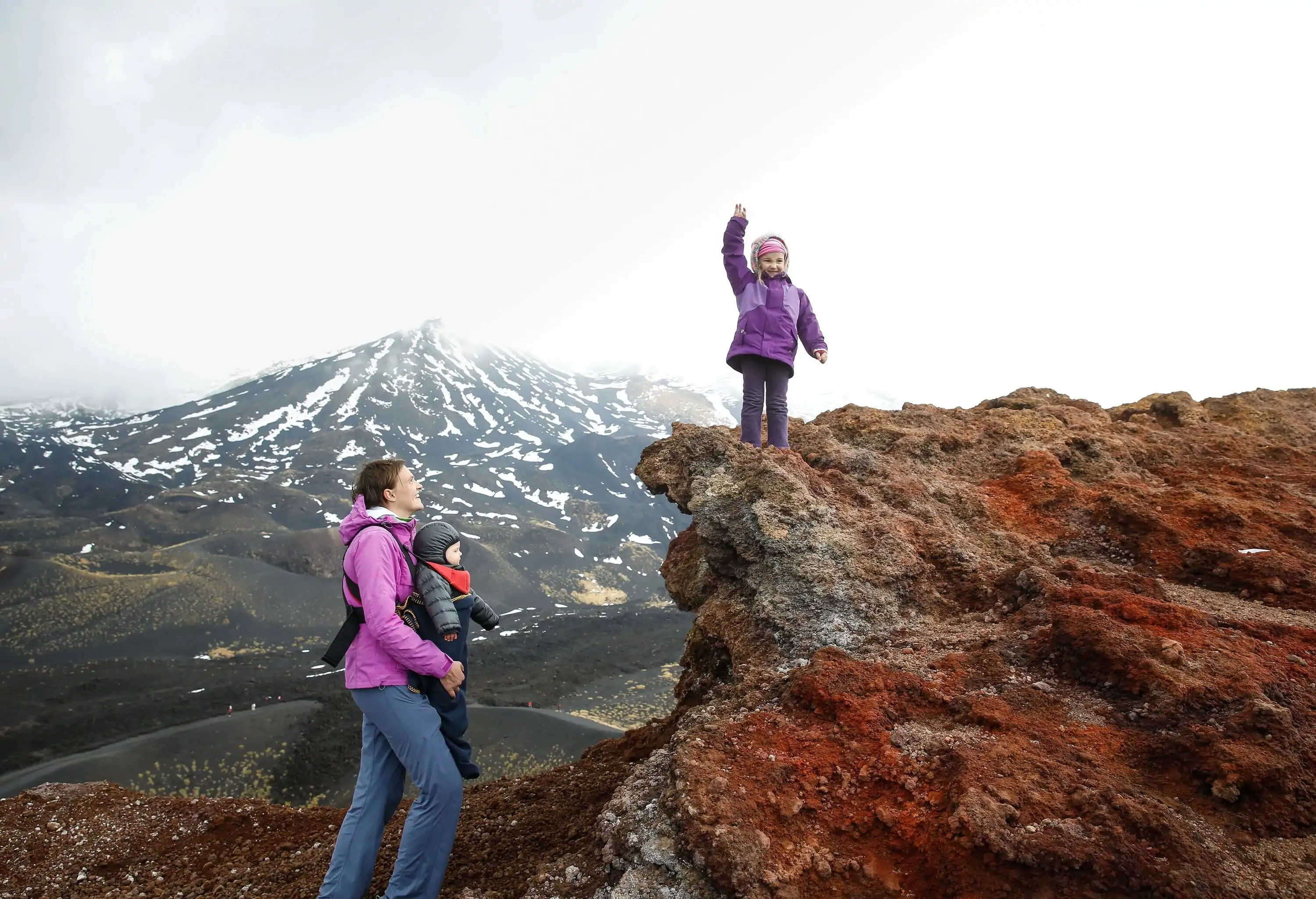 Mother with her baby and triumphant toddler on the edge of a crater on Mount Etna, volcano on the island of Sicily, Italy. Active family lifestyle, traveling family concept.