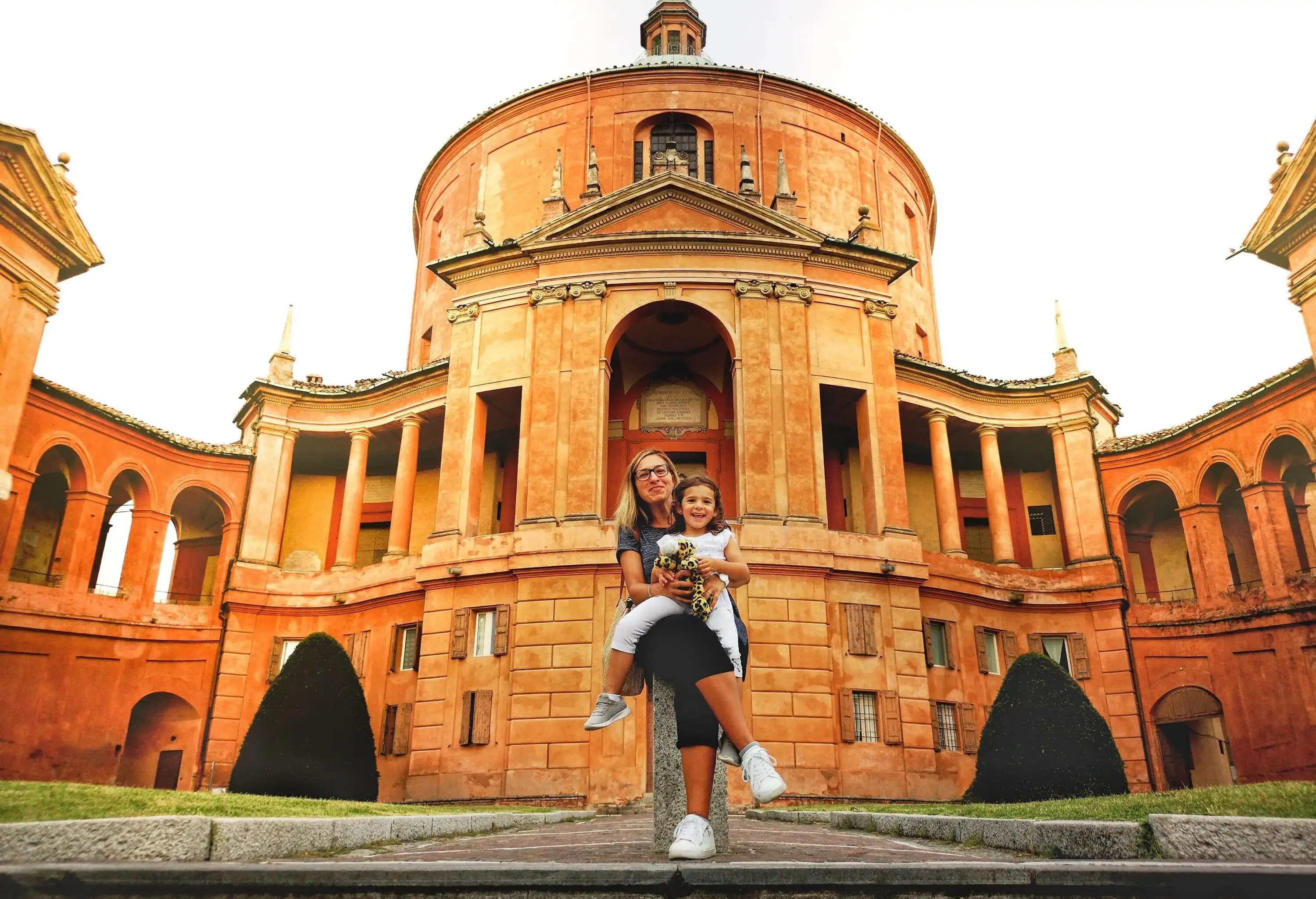 Woman sits on a concrete slab with a child on her lap in front of the Sanctuary of San Luca.