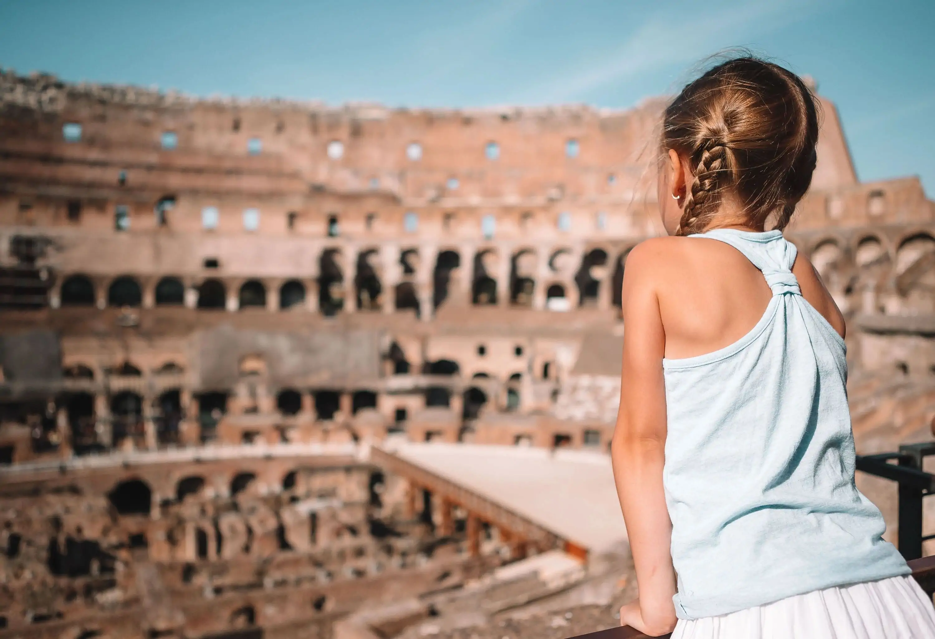 A little blonde girl is standing in an elevated area of the Roman Colosseum, looking at the surrounding.