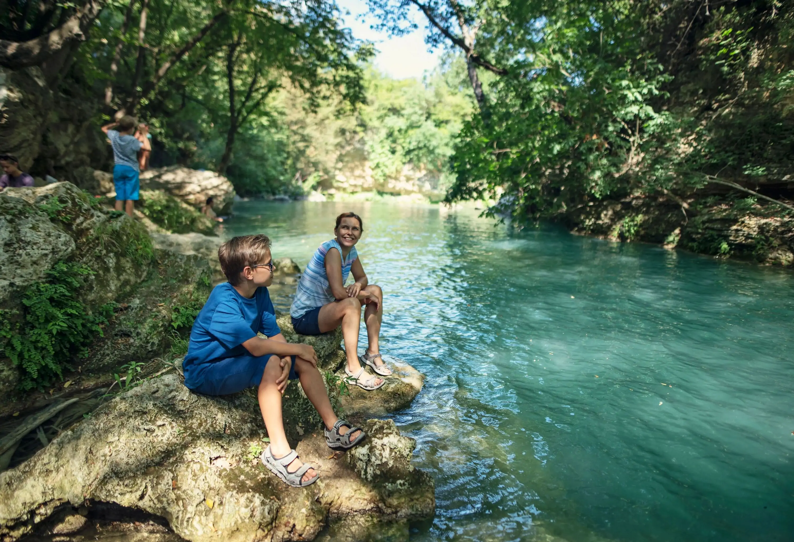 A mother and son sit on a riverside rock surrounded by tall green trees.
