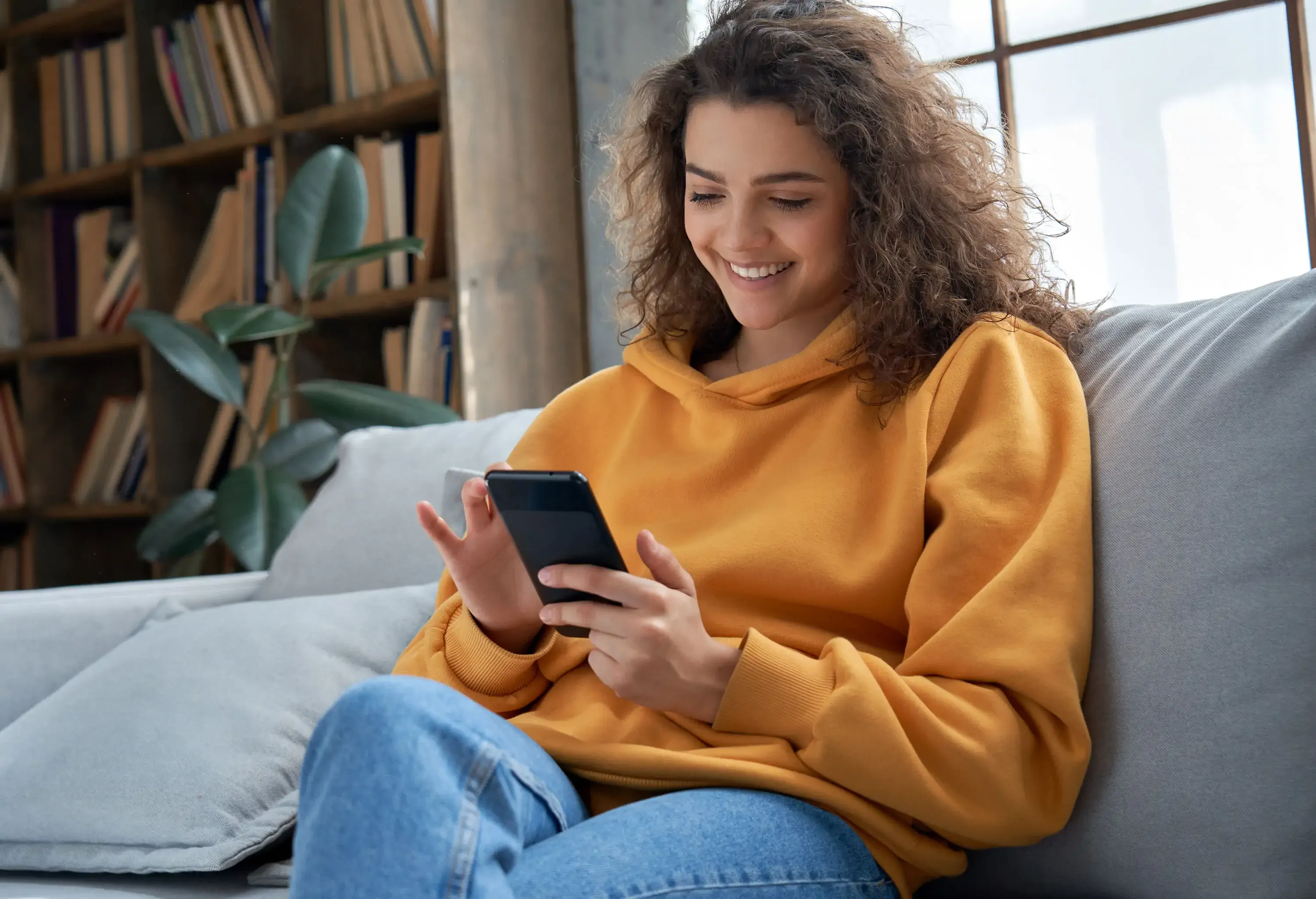 A woman in a hooded sweater sitting on a couch while browsing through her phone.
