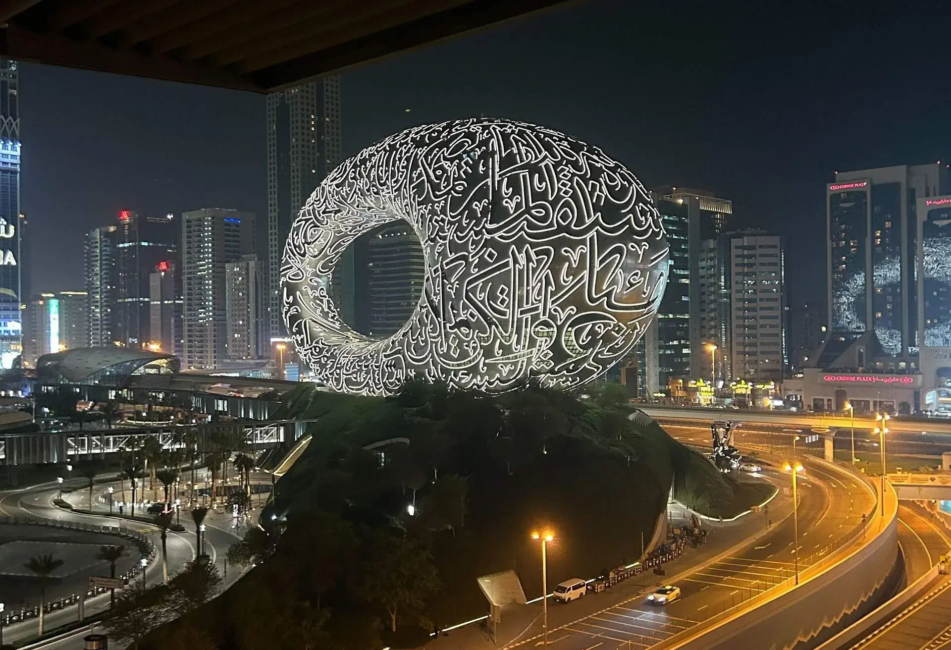 A beautiful monument in the shape of a sphere with Arabic letters written in bright LED lights stands next to a road.