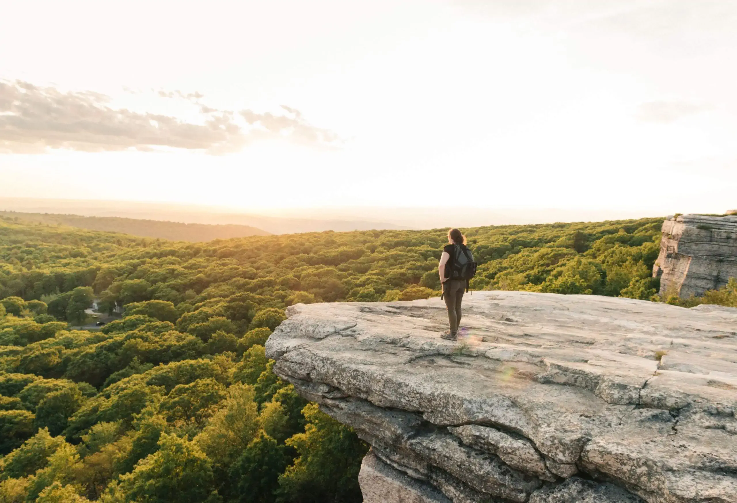 A person standing on a cliff's edge gazing to the valleys covered in thick forest.