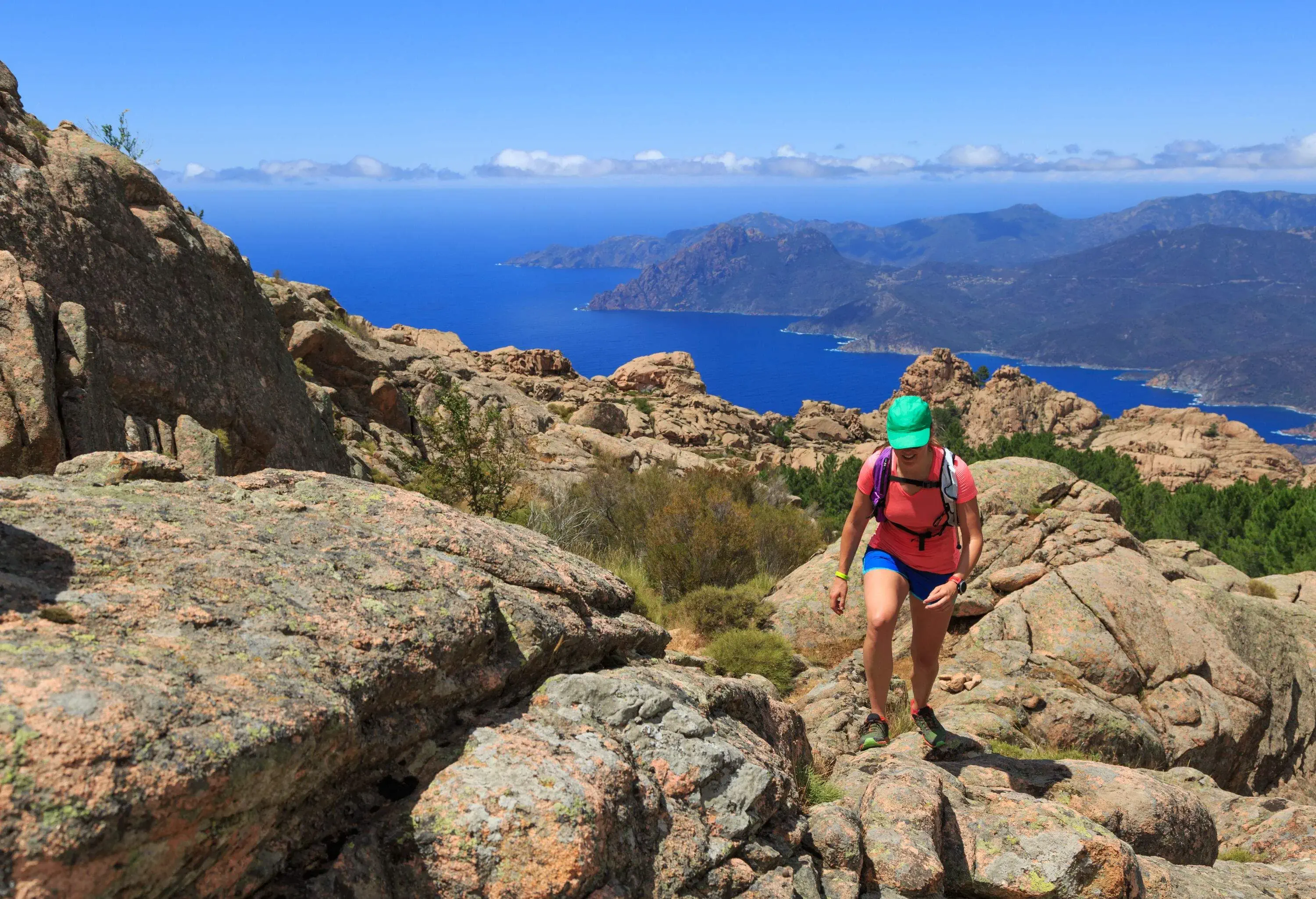 A woman in a pink shirt and hat hikes a rough mountain trail, with a view of the valleys and ocean in the distance.