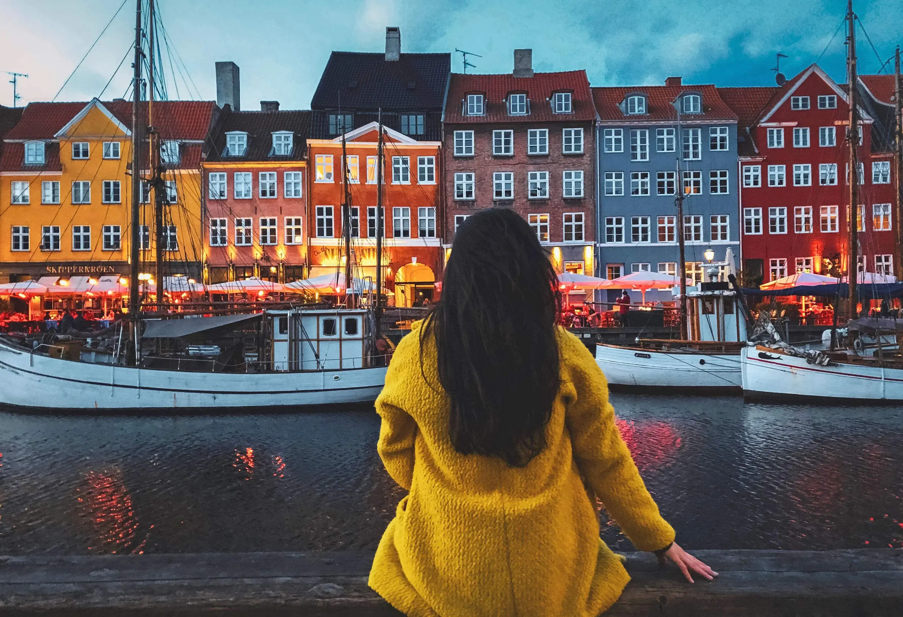 A person in a yellow cardigan is seated on a wooden rail, gazing out at the harbour and the boats moored there.