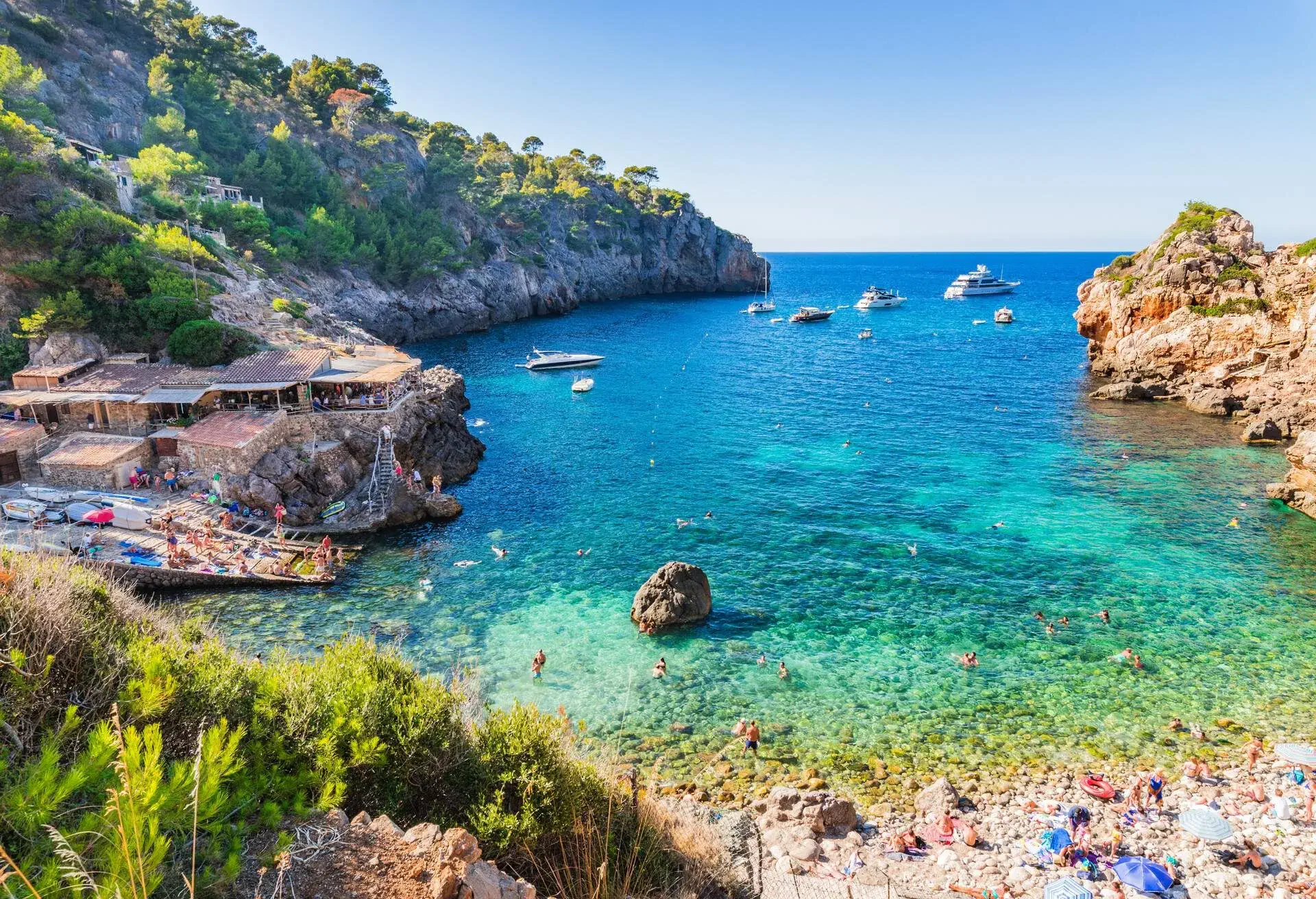 People lounging on a bay's rocky shoreline, with boats sailing in the distance.