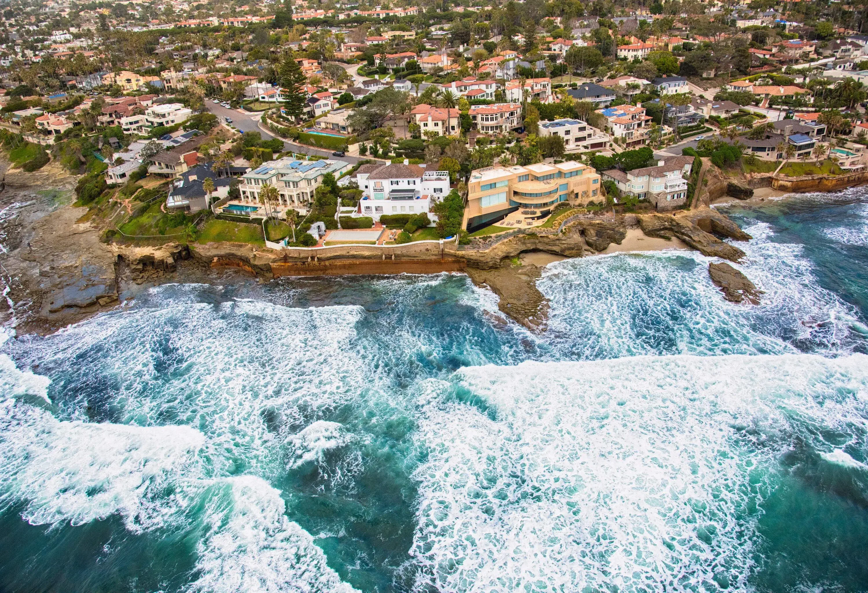 White sea waves crash into the rocky, hilly coast with colourful houses.