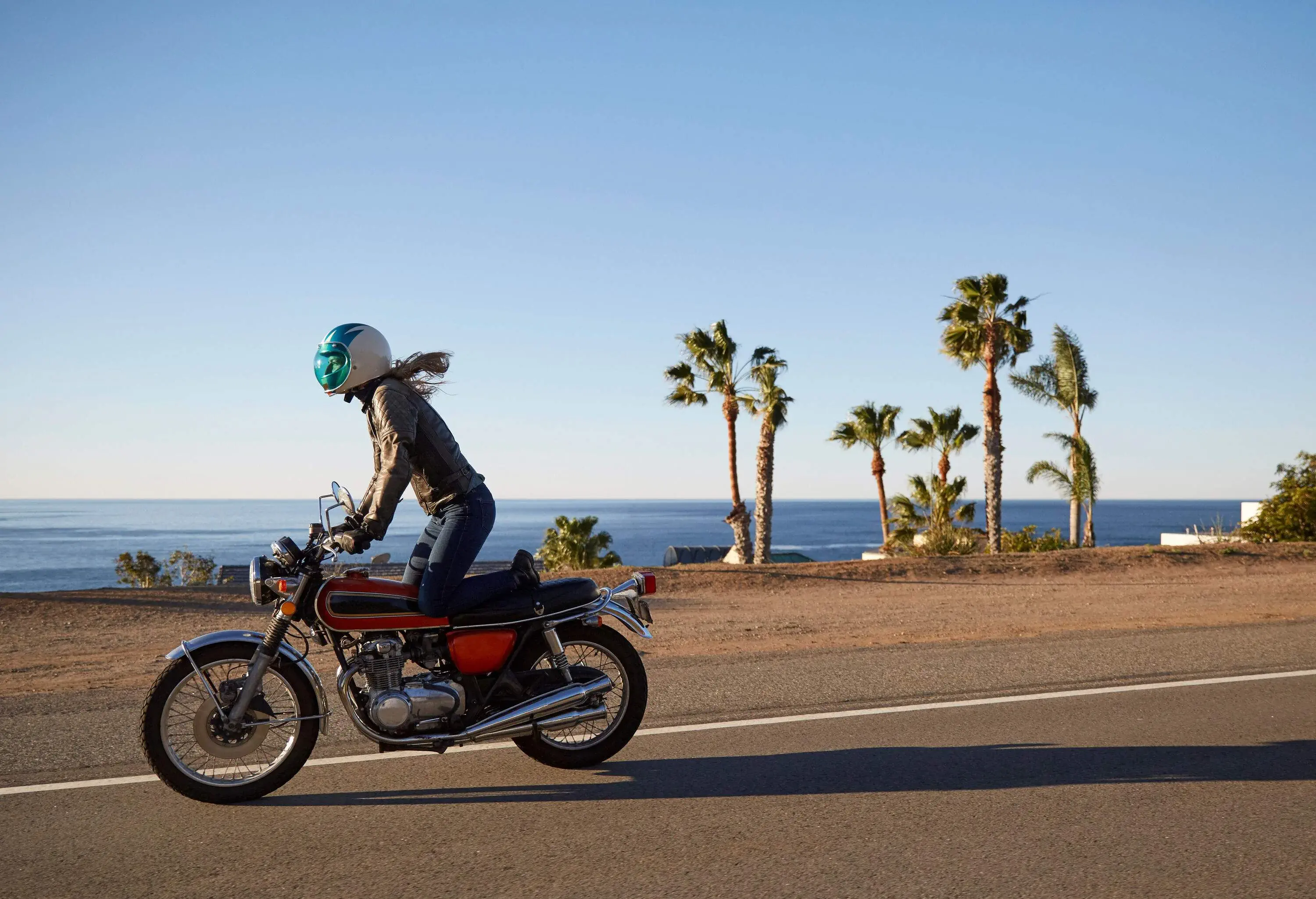 A woman in motor riding gear enjoys riding a motorbike along a paved road that runs alongside the captivating sea.