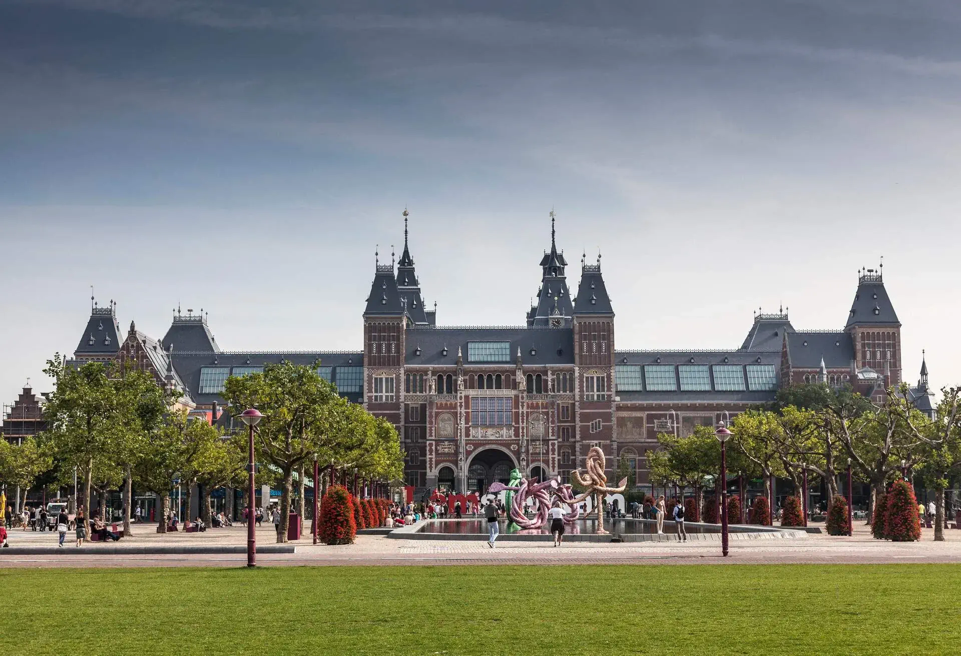 Gothic architecture and a palatial museum façade with rows of tall green trees and a fountain pond with a large rope-like structure in the middle.