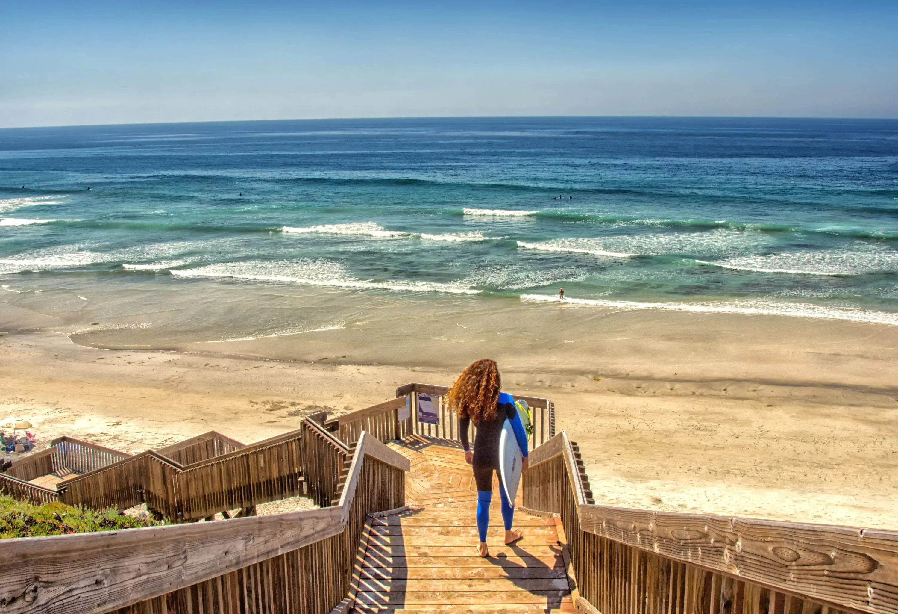 View of woman wearing wetsuit and holding surfboard looking at the sea from wooden staircase on a beach