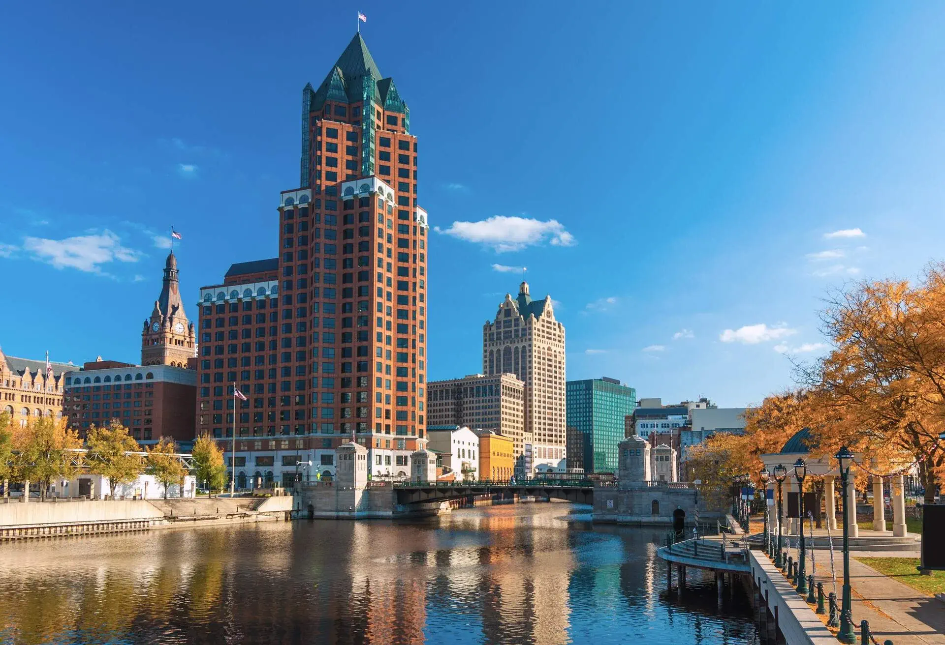 Downtown Milwaukee skyscrapers (prominently showing Milwaukee Center) with the Milwaukee River and orange colored trees during Autumn in the foreground.