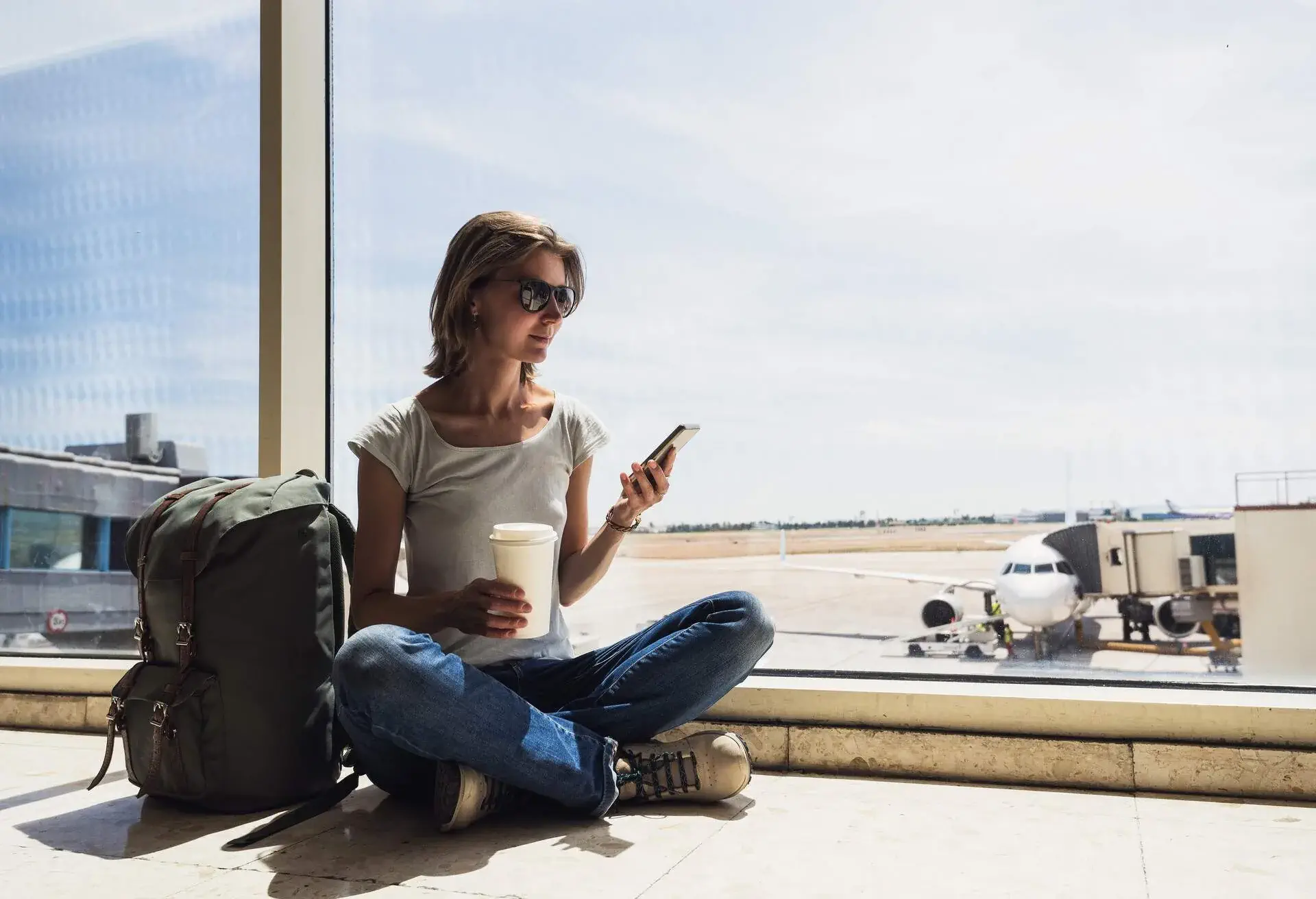 A young girl eagerly awaiting her plane at the airport, comfortably seated on the floor, holding a drink and her phone in her hands, framed by the transparent glass wall of the boarding area.