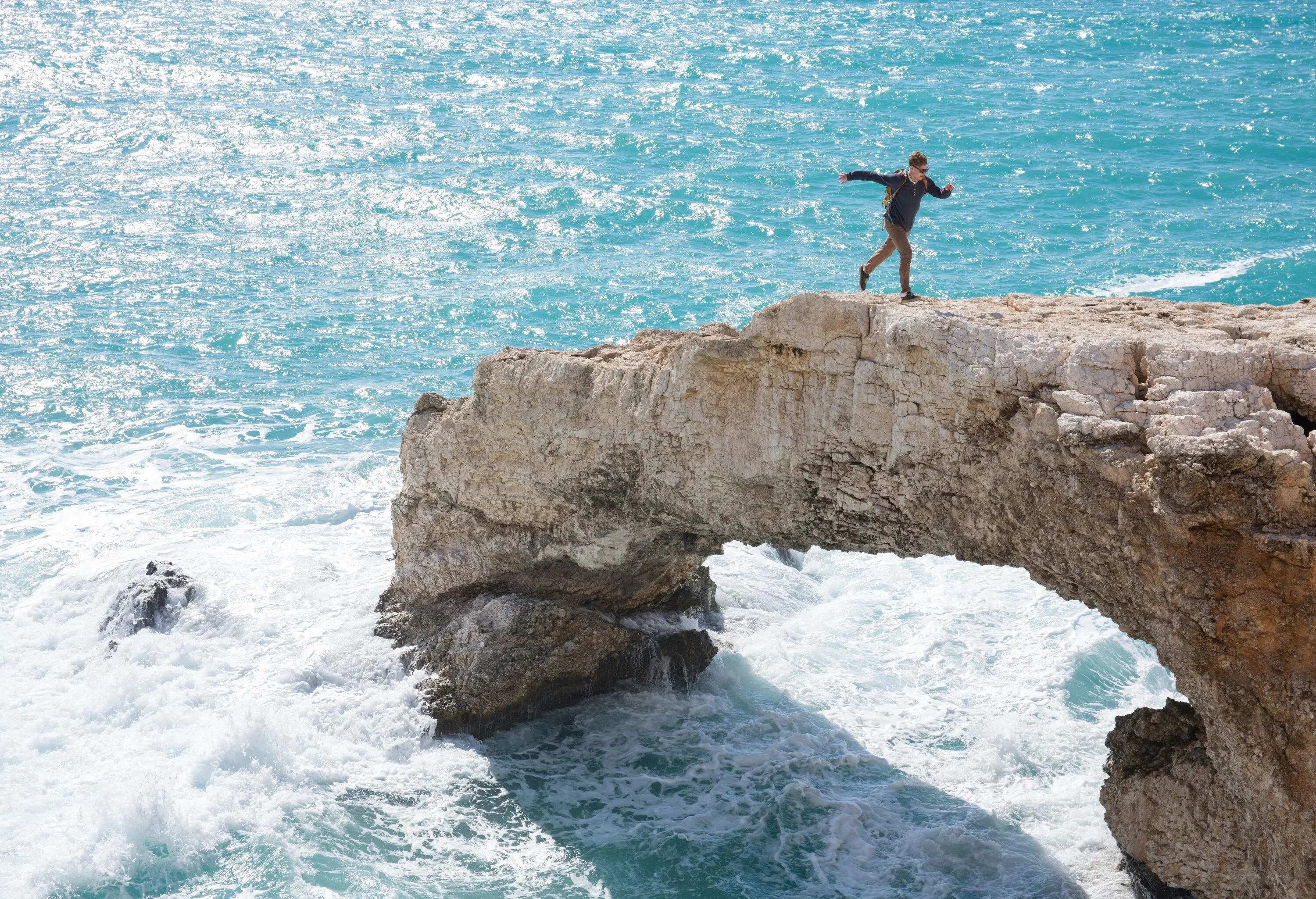 A man runs on top of an arch rock formation above the crashing waves of an ocean.