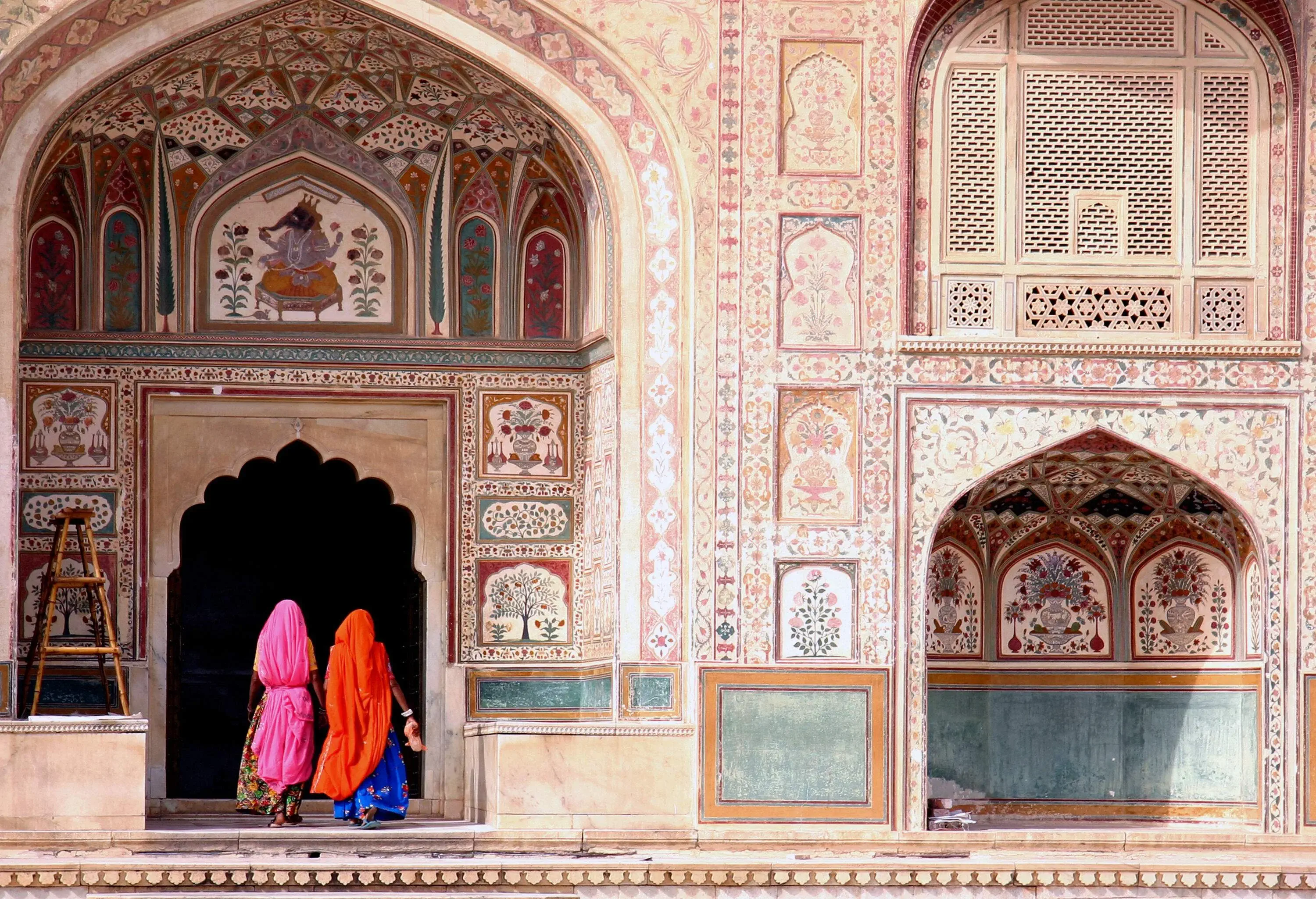 Two women gracefully wrapped in colourful linen walk along a pointed arch walkway, bordered by walls adorned with intricate carvings.