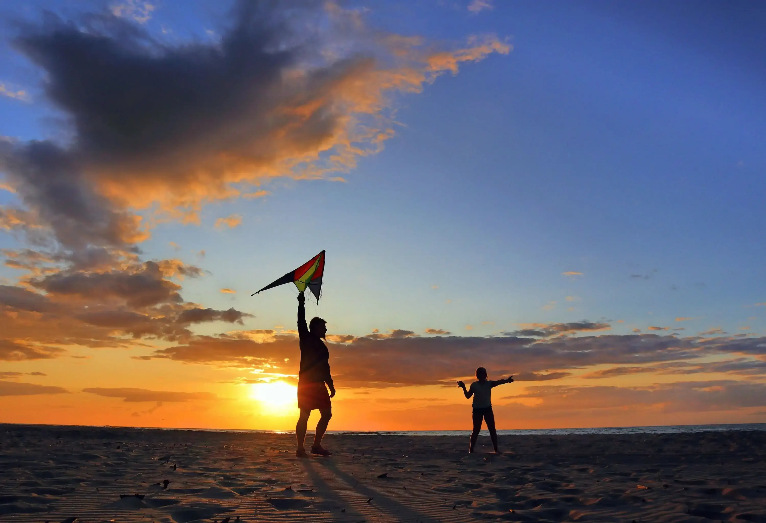 As the sun sets over the stunning beach, a charming couple can be seen joyfully attempting to fly a colourful kite, their elegant silhouettes standing out against the vibrant hues of the sky.