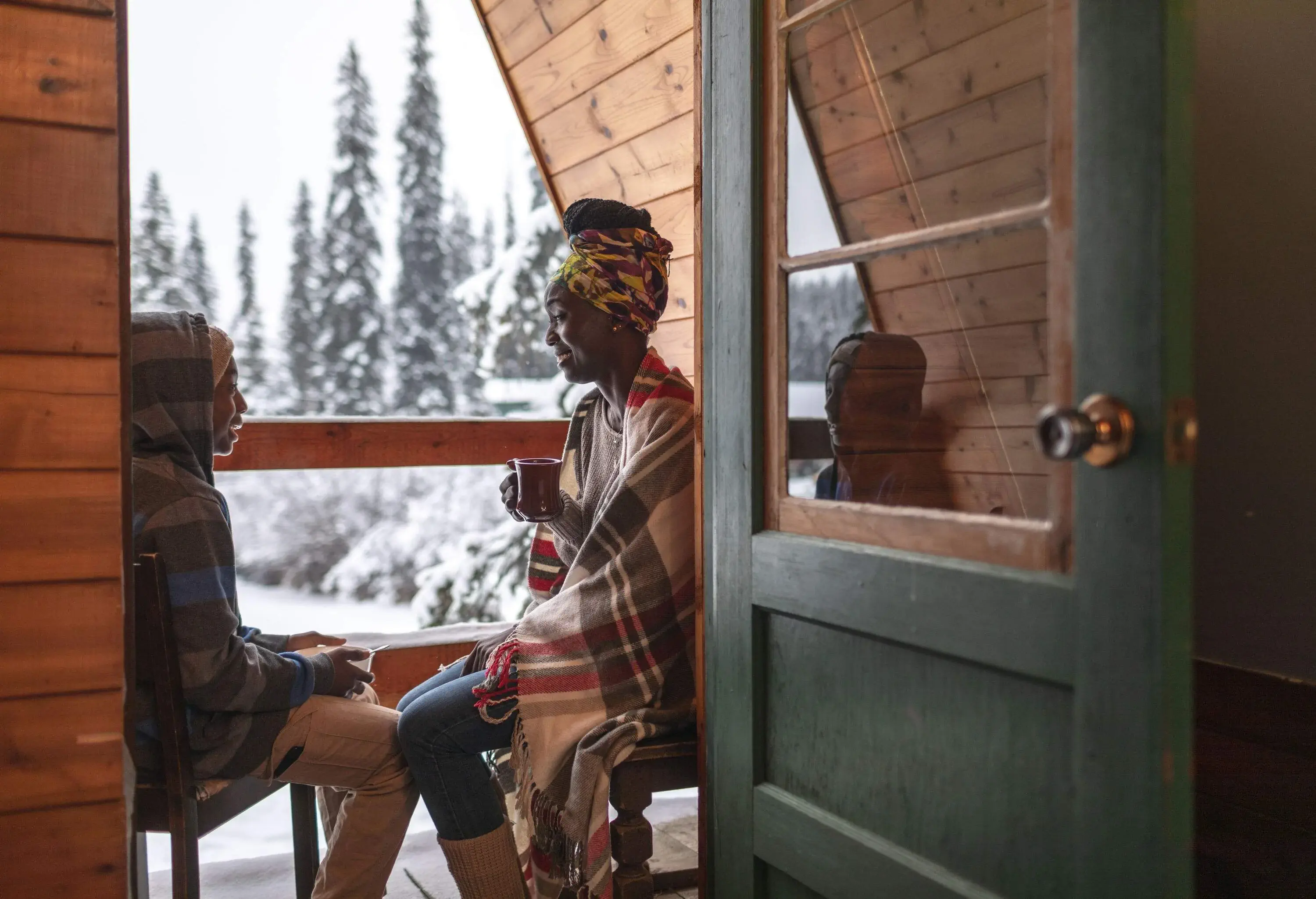 A mother and son sitting and talking on a small balcony with warm drinks in their hands.