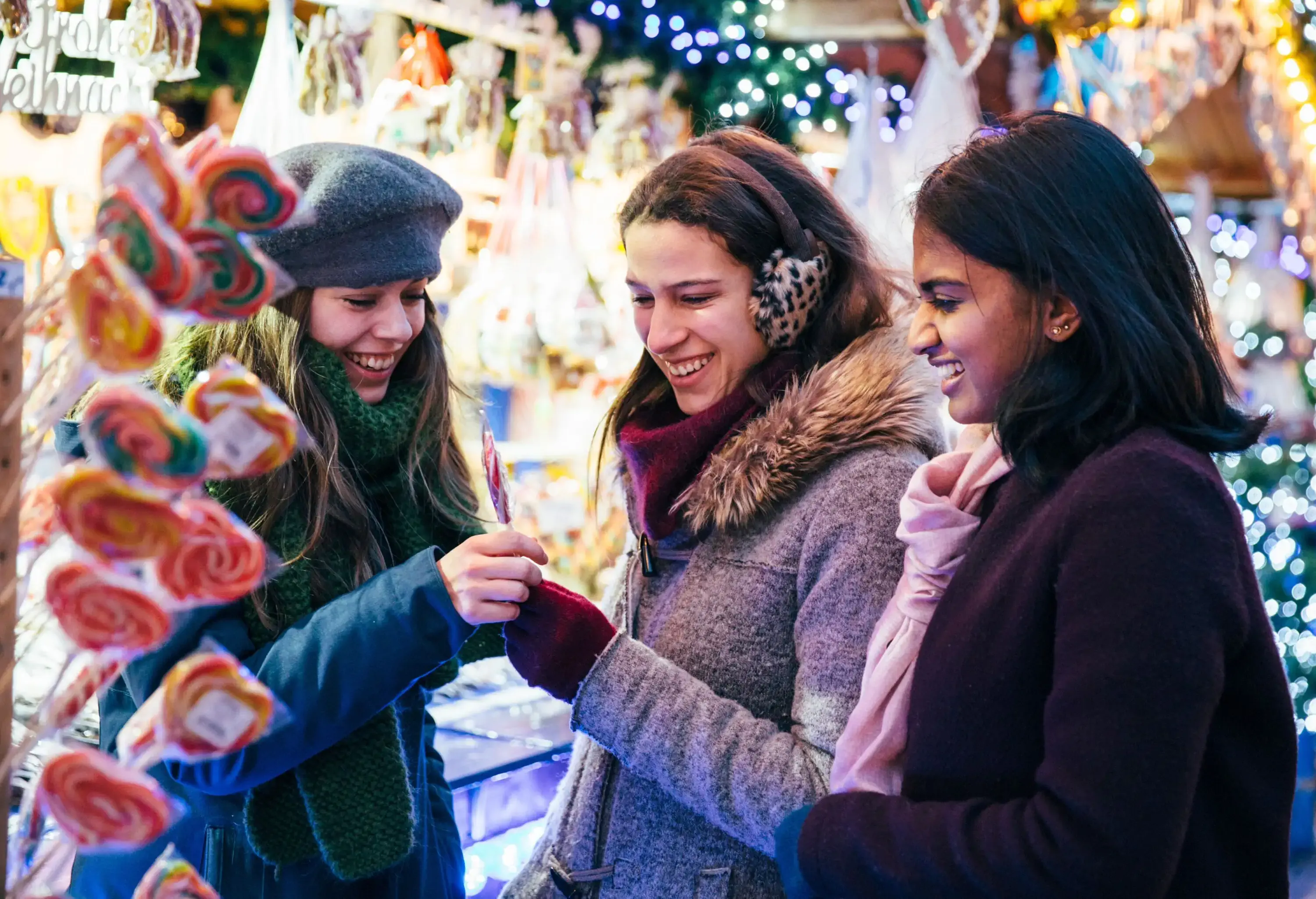 Three smiling female friends wearing winter clothes shopping at Christmas market