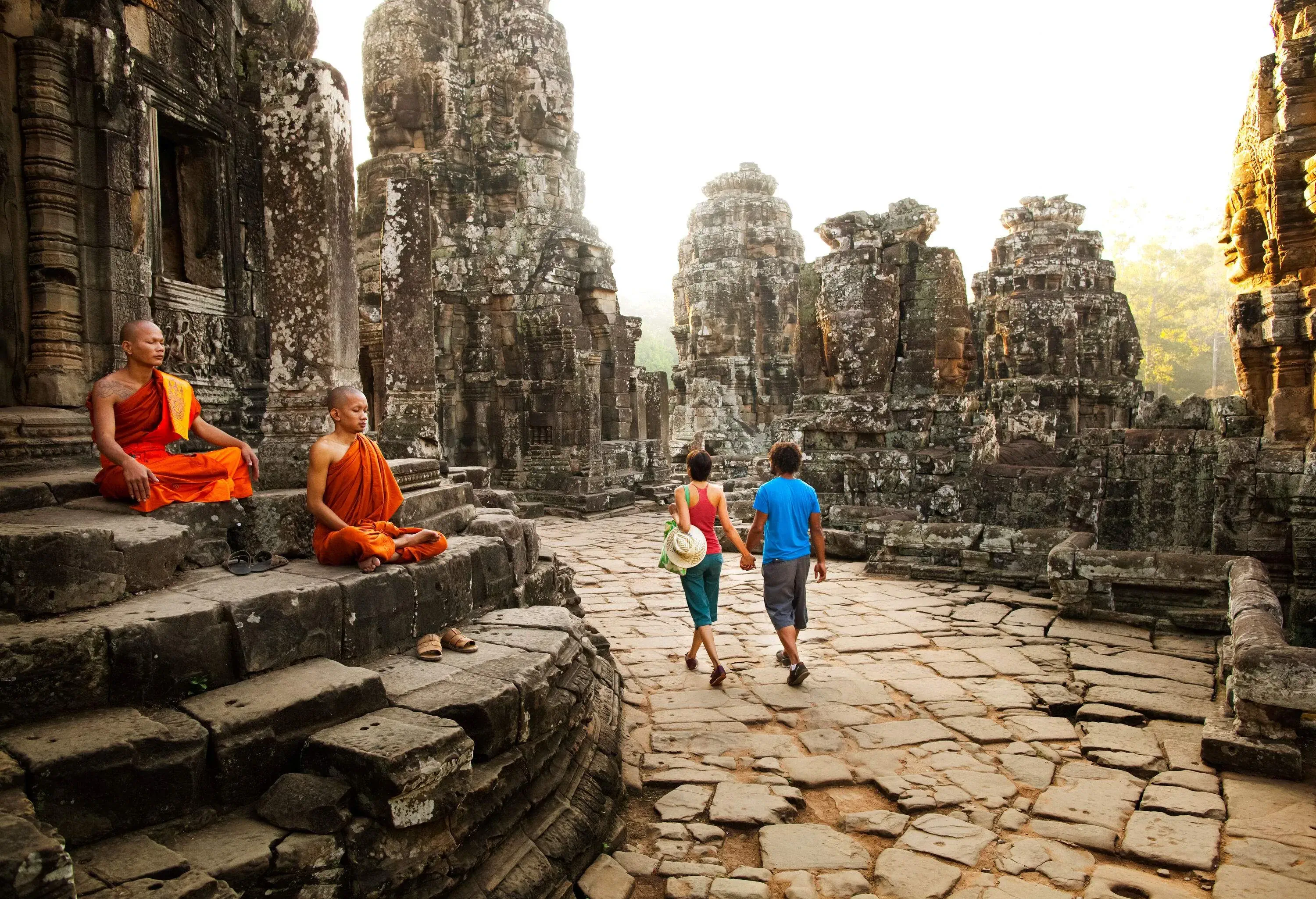 A couple in casual clothes holding hands as they walk past an ancient temple staircase with two monks meditating.