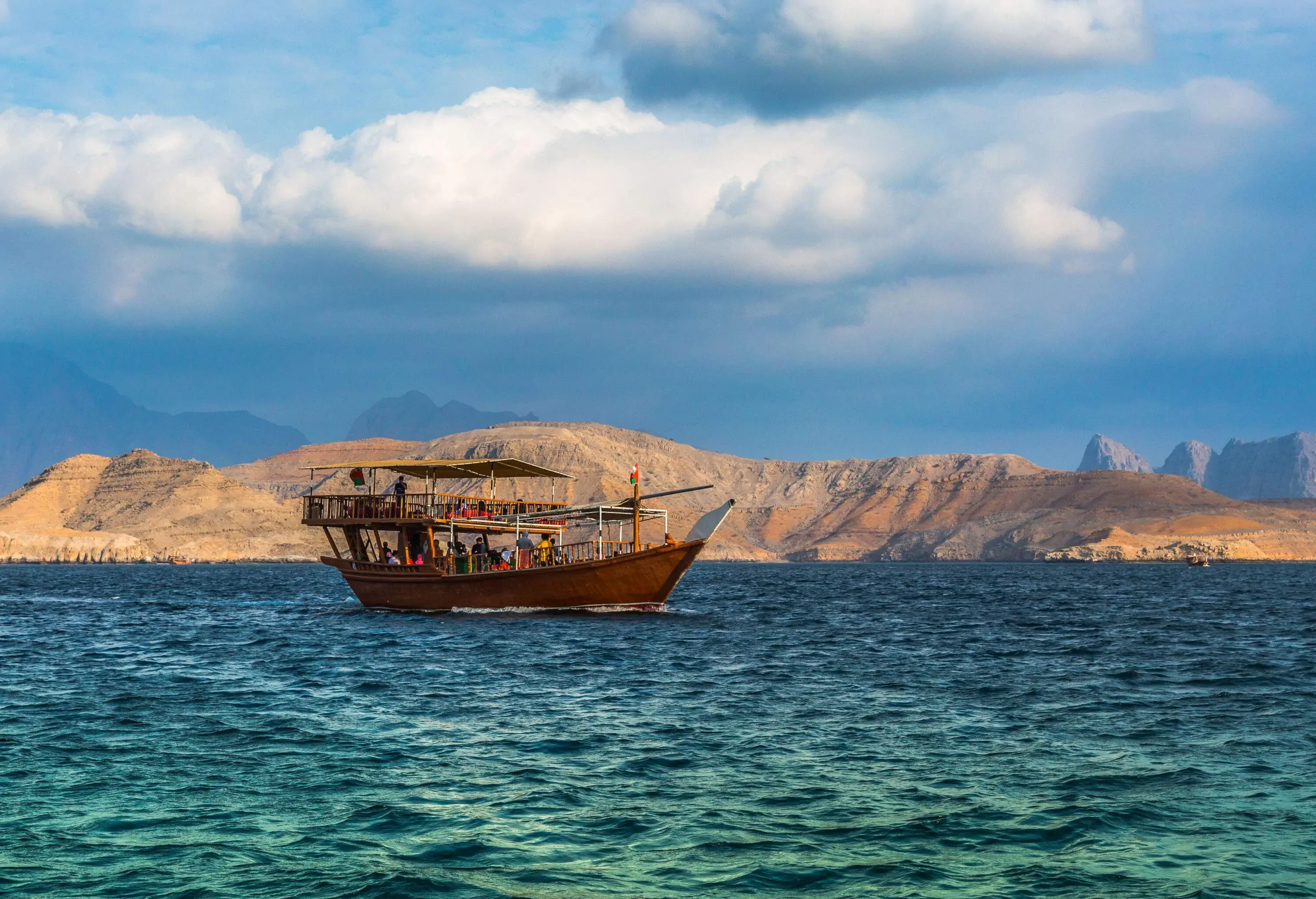 A dhow boat carrying passengers cruising in the sea, with sandy mountains in the background.