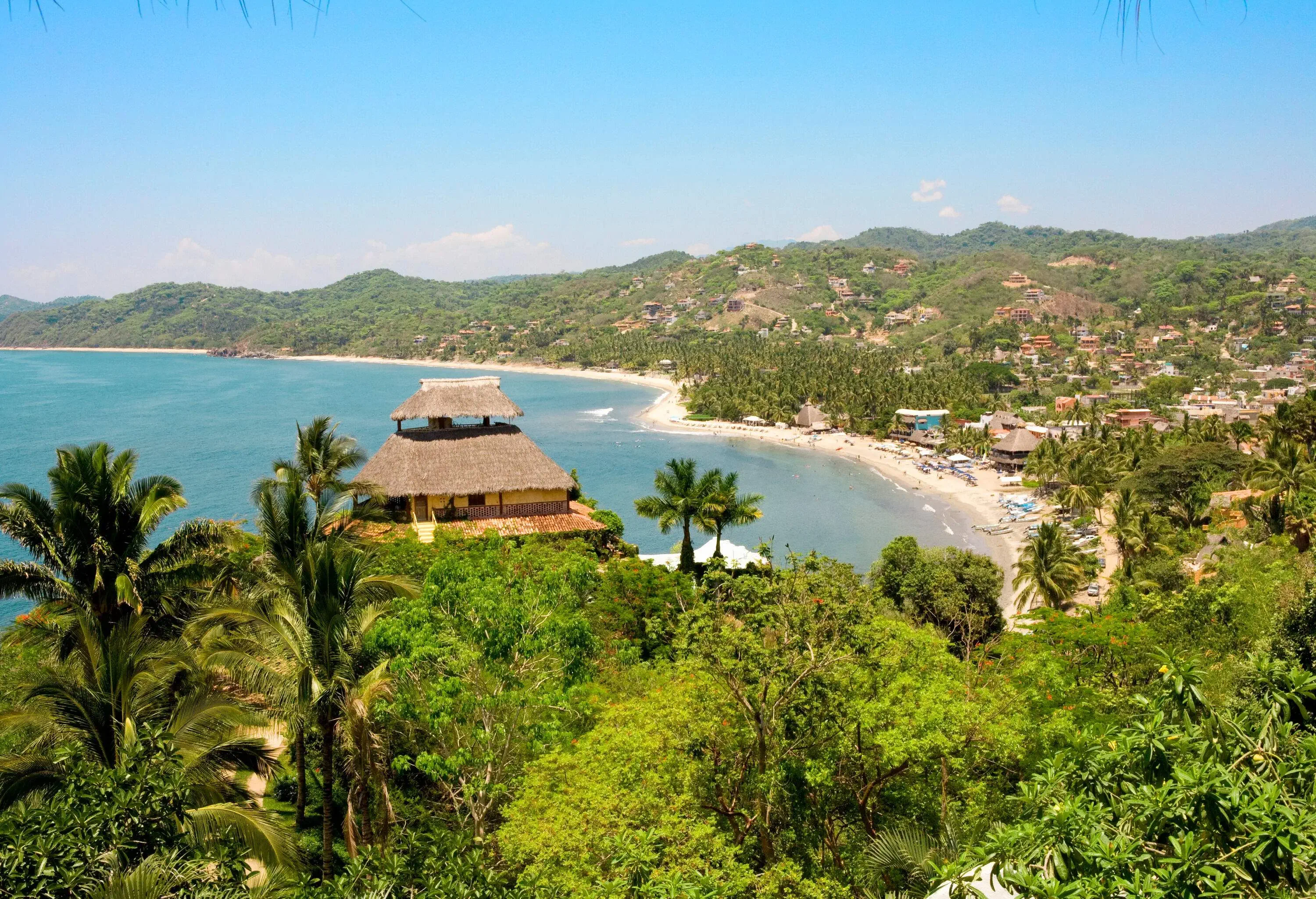 A quaint village perched on a slope overlooks a tranquil bay, surrounded by lush wooded areas and verdant rolling hills in the distance.