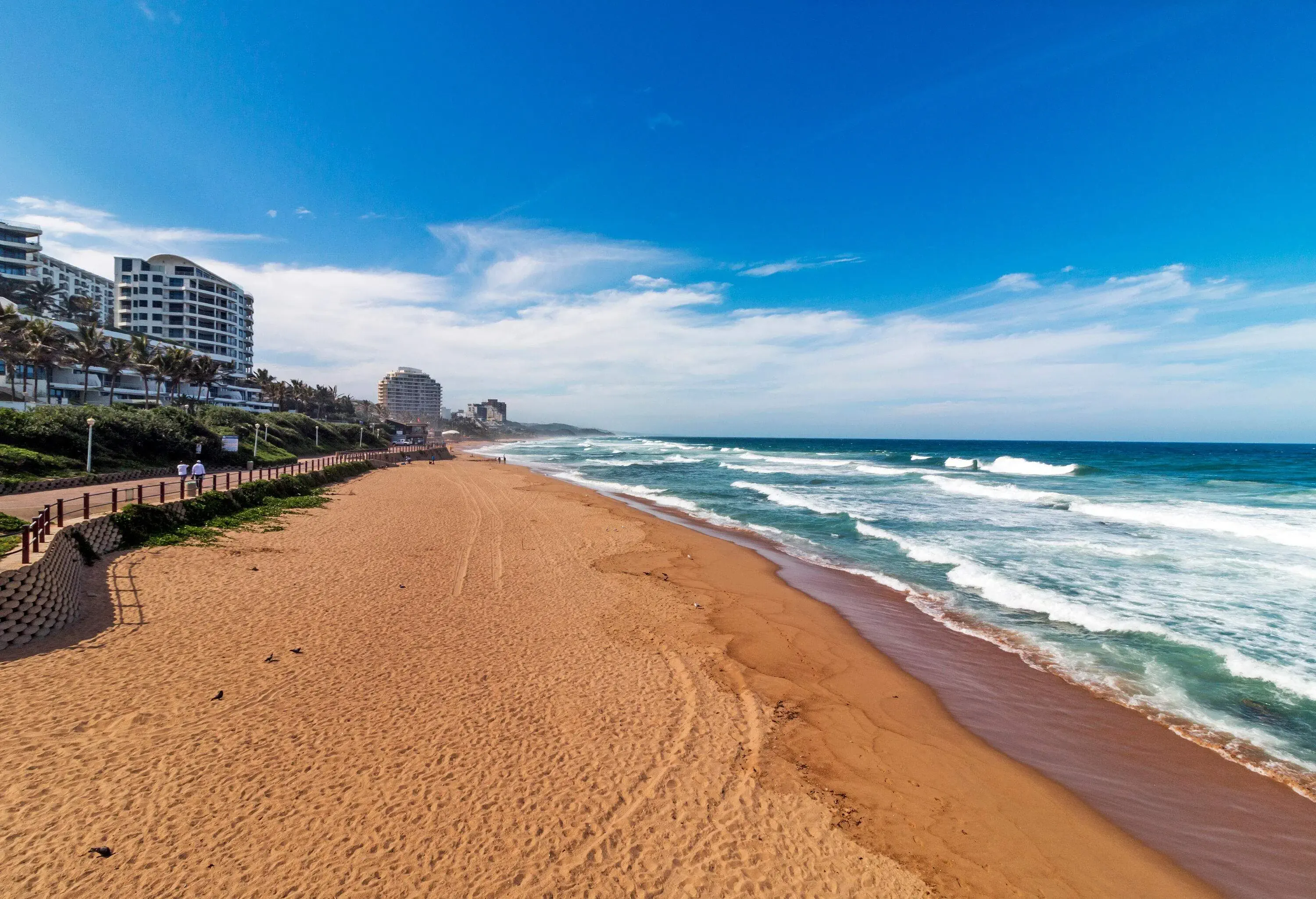 An empty beach with rough waves along a promenade garden and waterfront buildings.