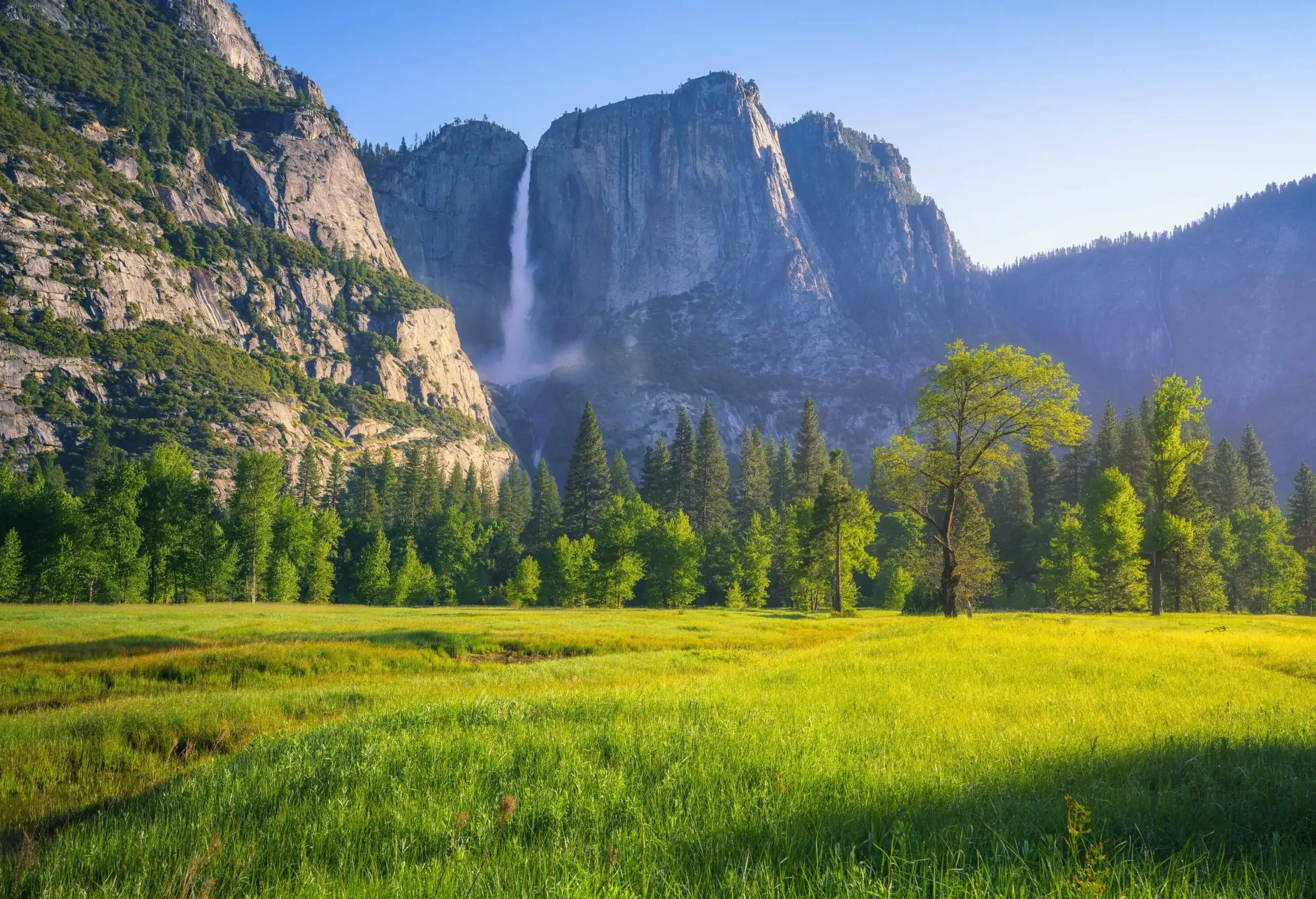 A verdant valley surrounded by tall green trees beneath the ragged mountain range with a waterfall.