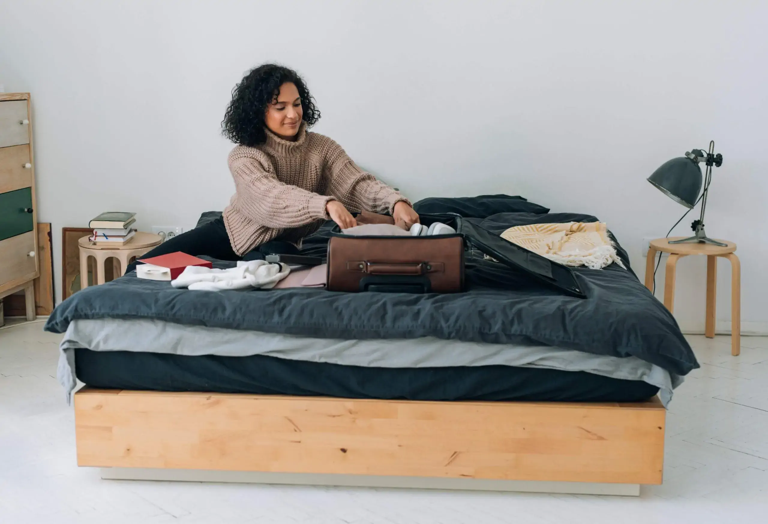 A curly-haired lady fixing her clothes into a brown suitcase on the bed.