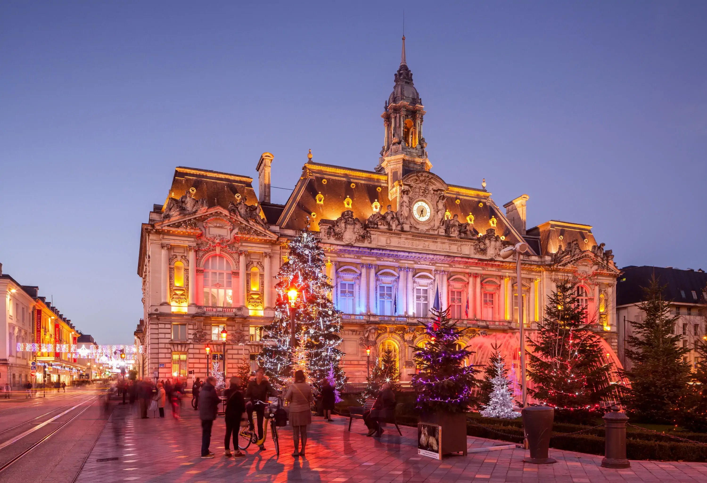 An exquisite Renaissance-style building shining in different colours with numerous illuminated Christmas trees in its front yard.