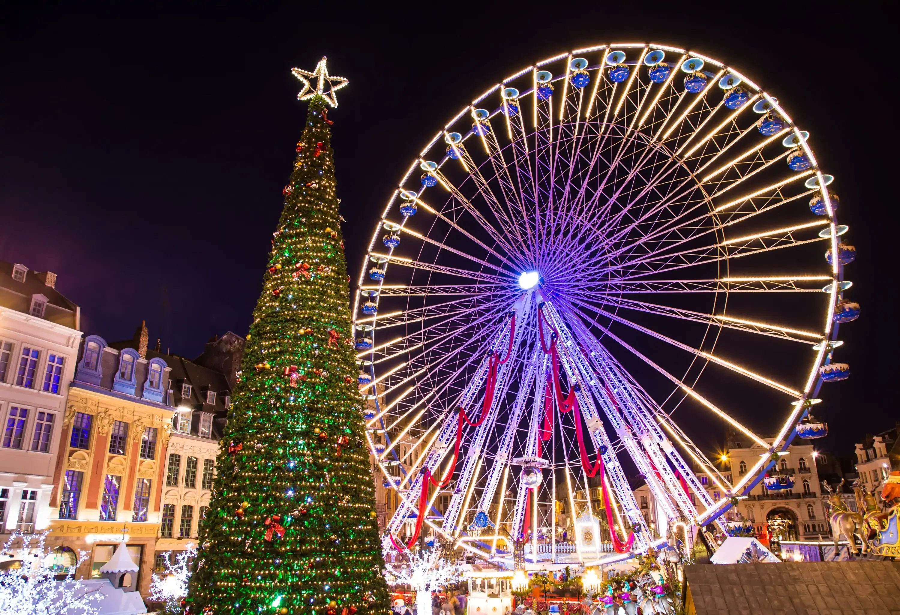 A festive nighttime scene featuring a brightly lit Ferris wheel and a decorated Christmas tree with a star on top, set in a European-style square during holiday celebrations.