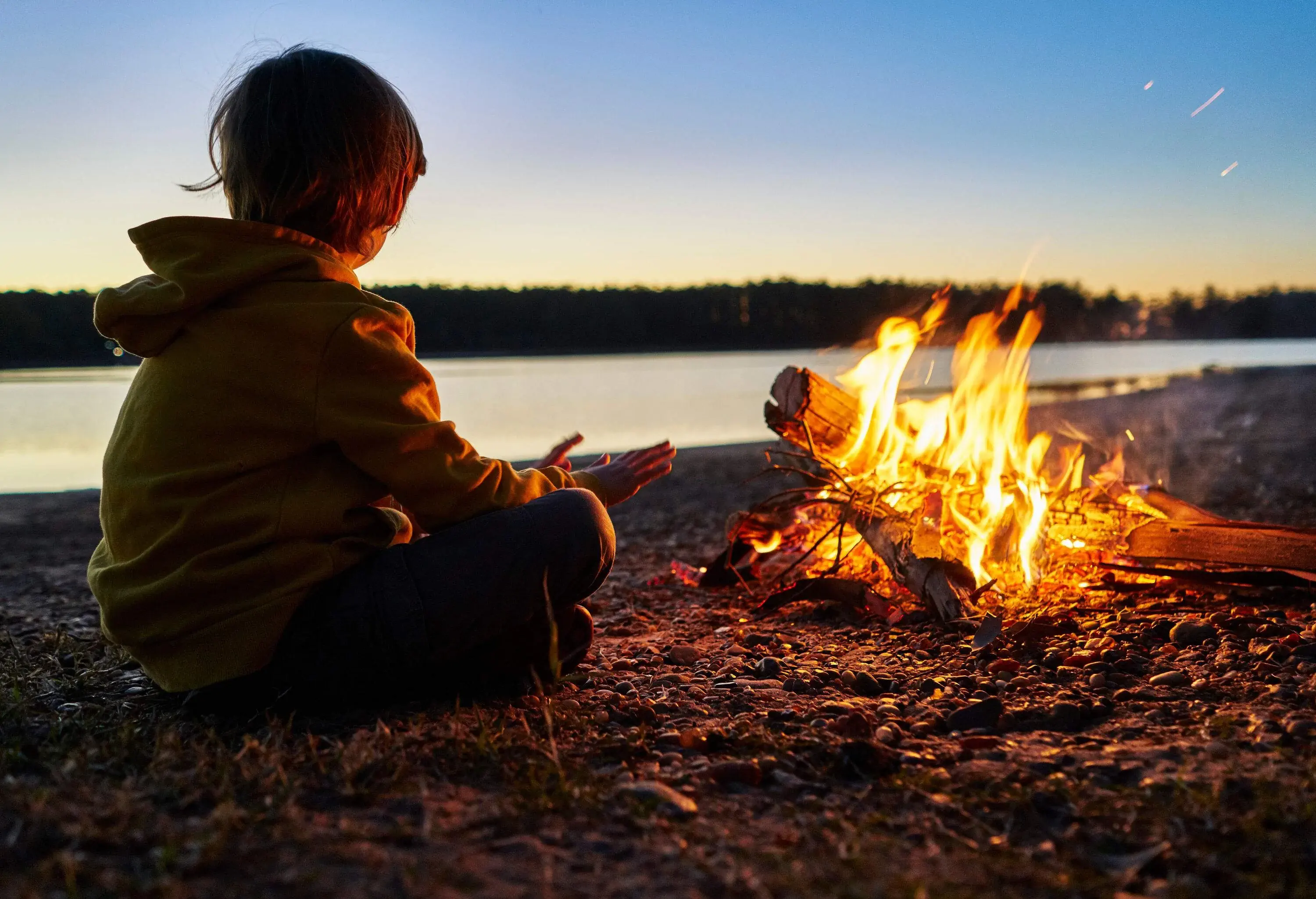 A child in a hooded jacket sits cross-legged in front of a bonfire with open hands.