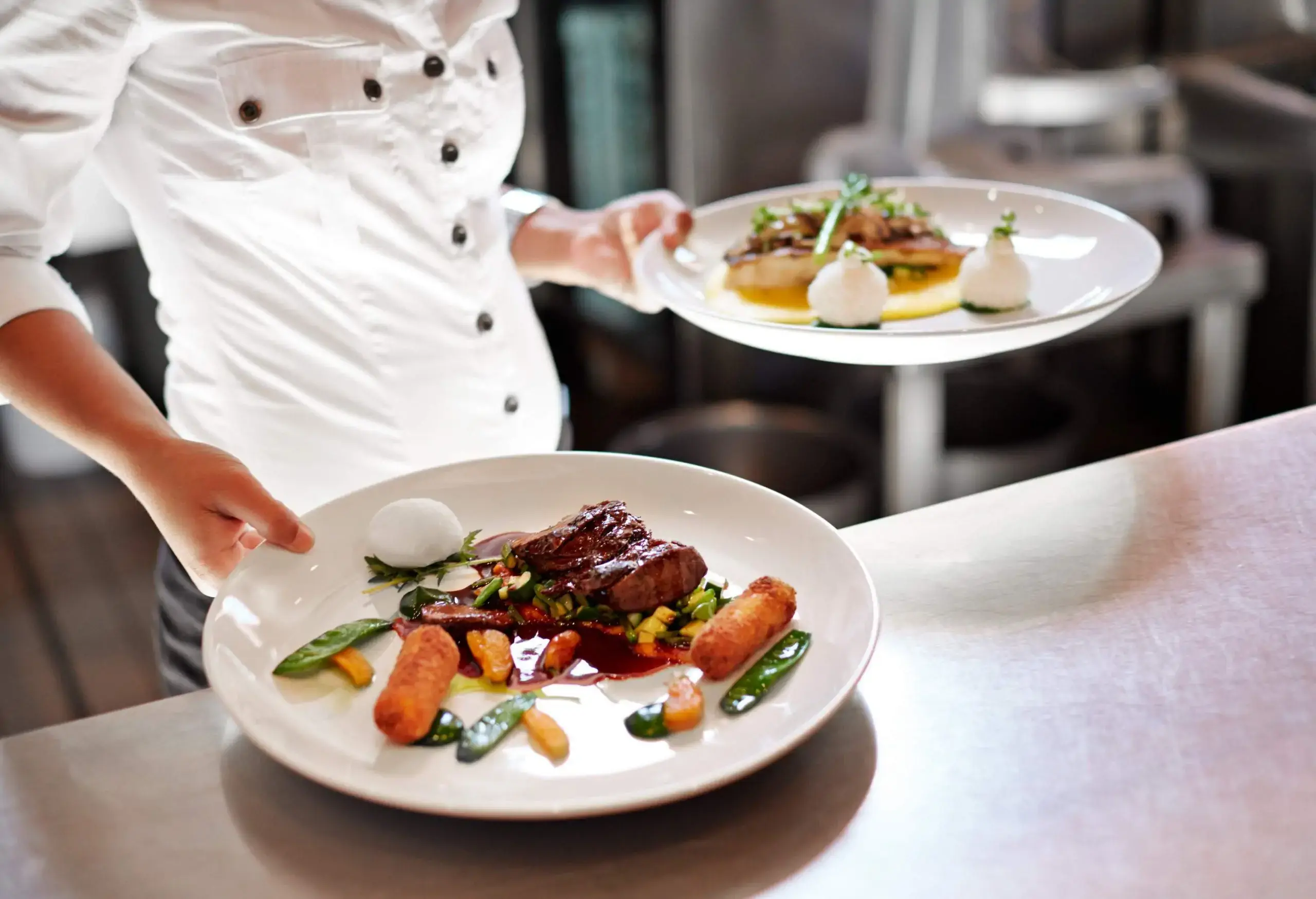 A woman picking up two plates from a stainless steel work table in a restaurant kitchen.