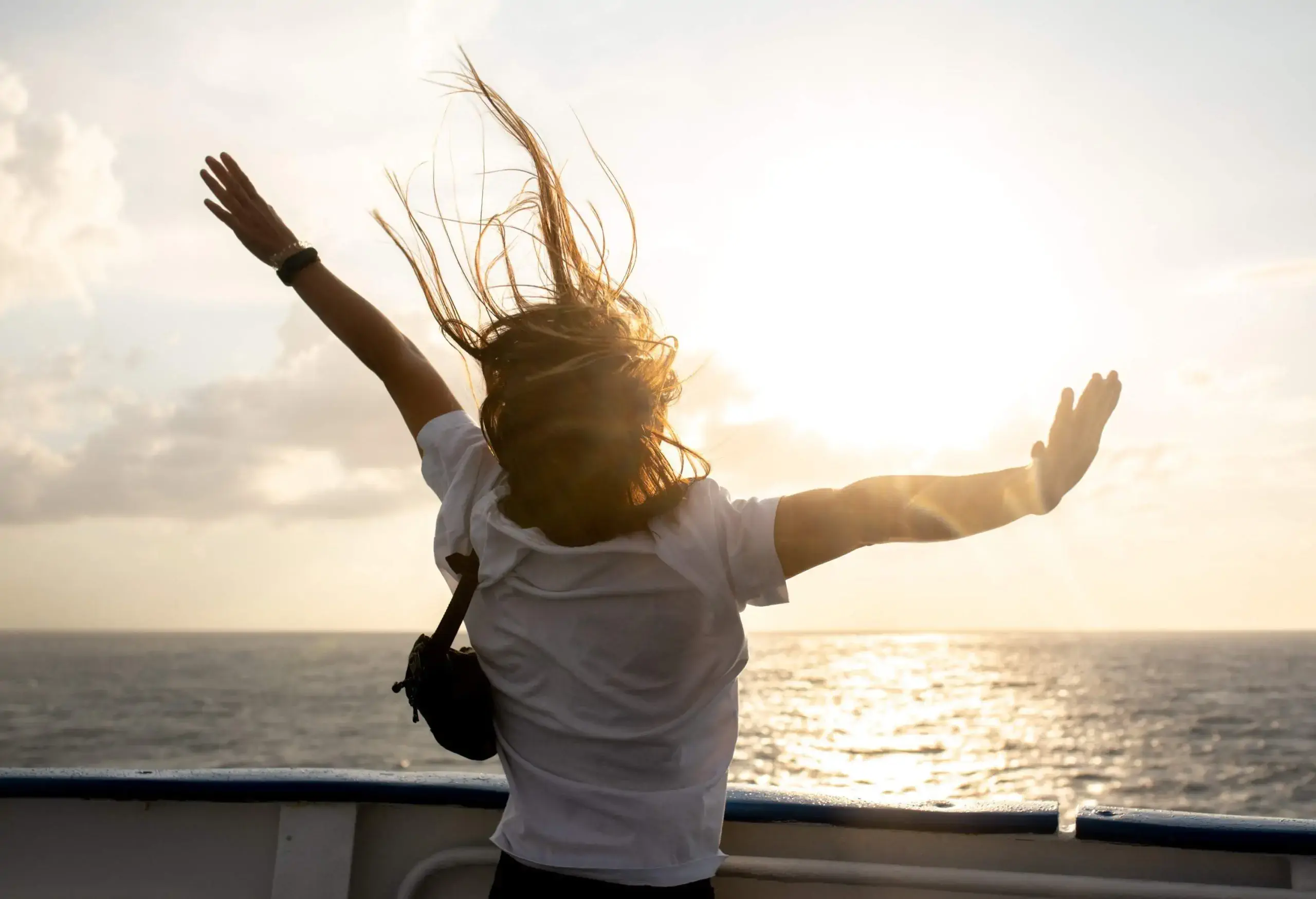 A woman spreads her arms as she stands on a ship's deck facing the sun above a vast ocean.