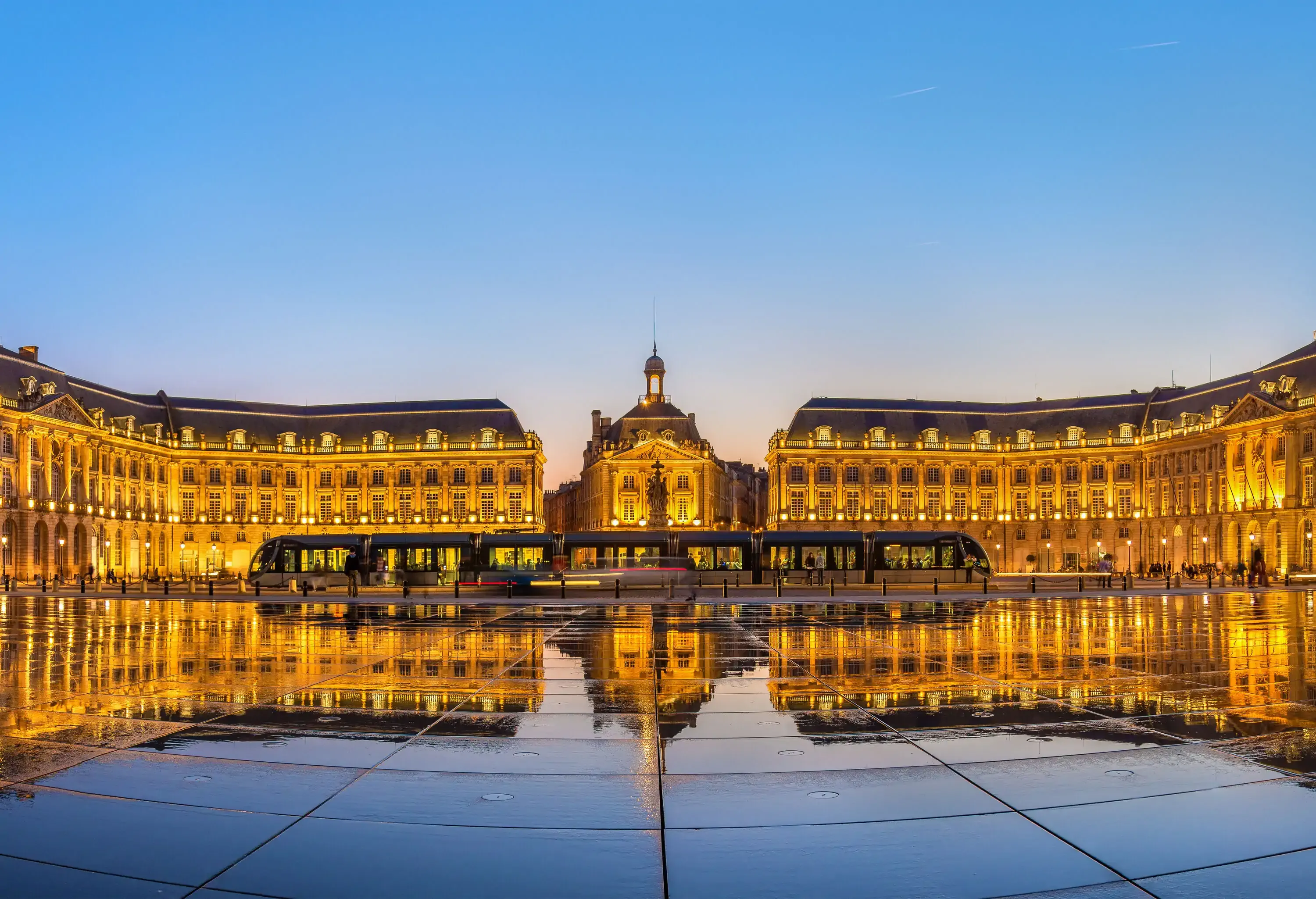 A tram passing by the magnificent Place de la Bourse, which is illuminated and mirrored in the water fountain on the ground.