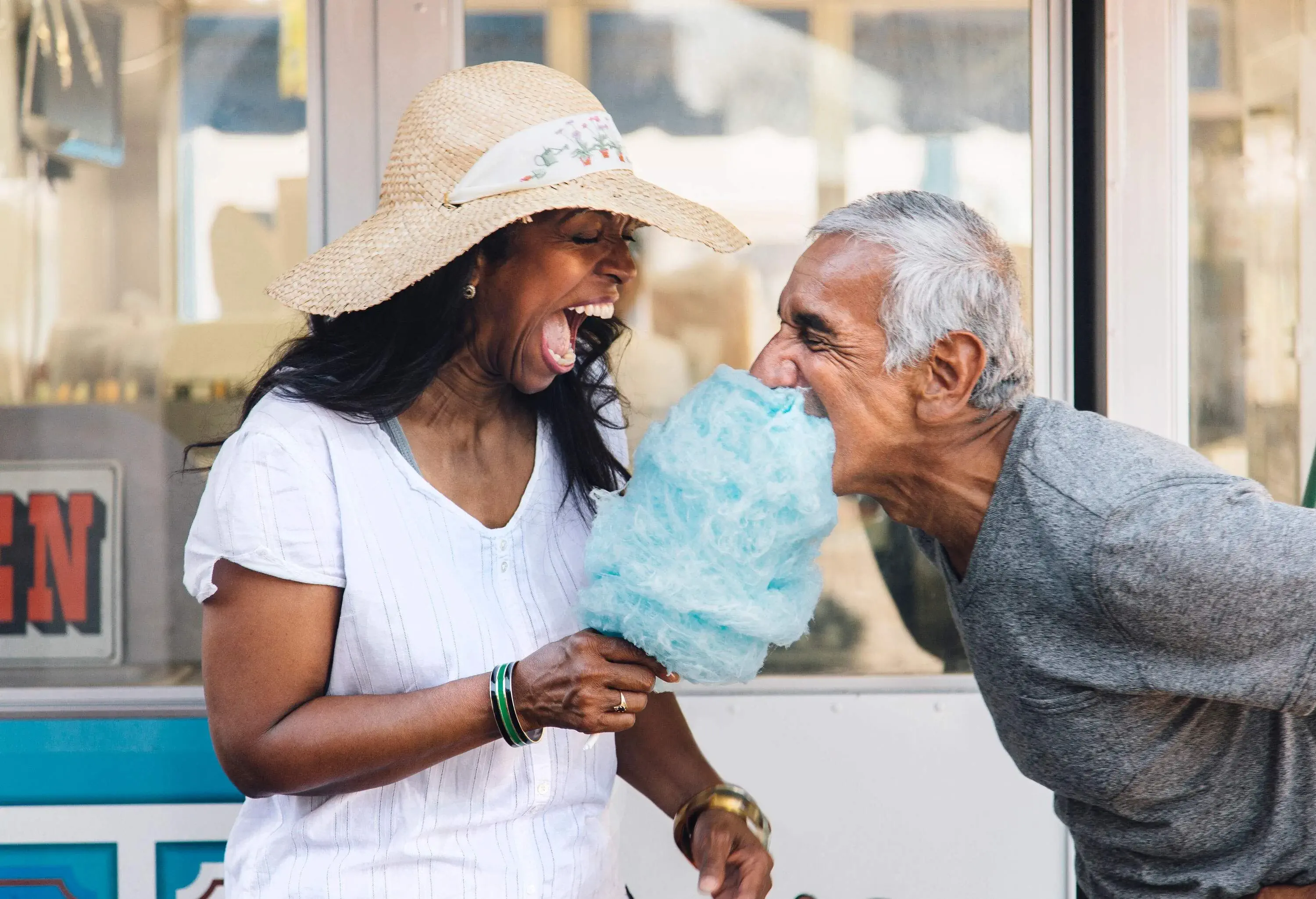 A woman with a wide open mouth holds a stick of cotton candy, and a man takes a big bite.