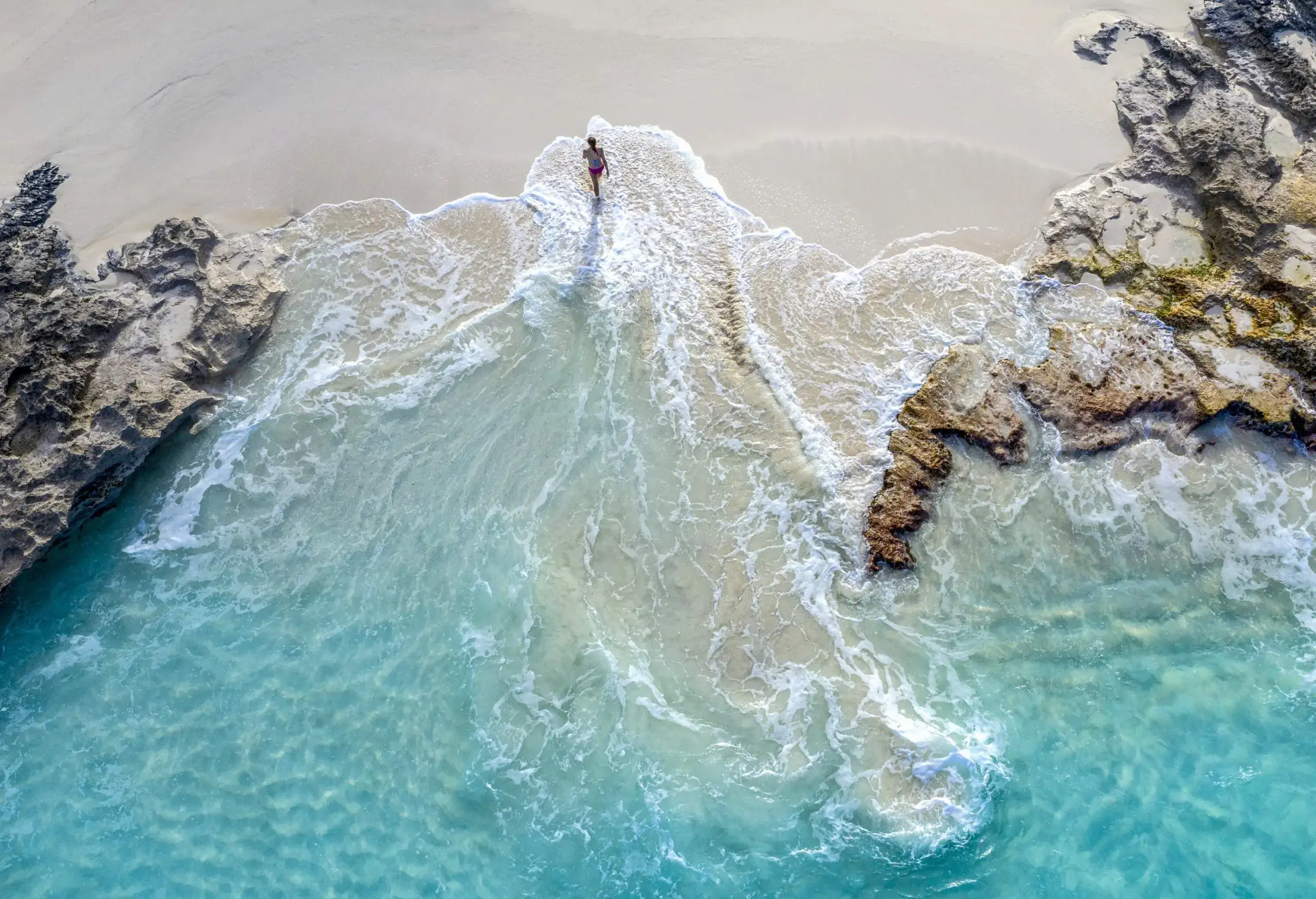 A person walks away from a beach with turquoise waves and rugged coast.