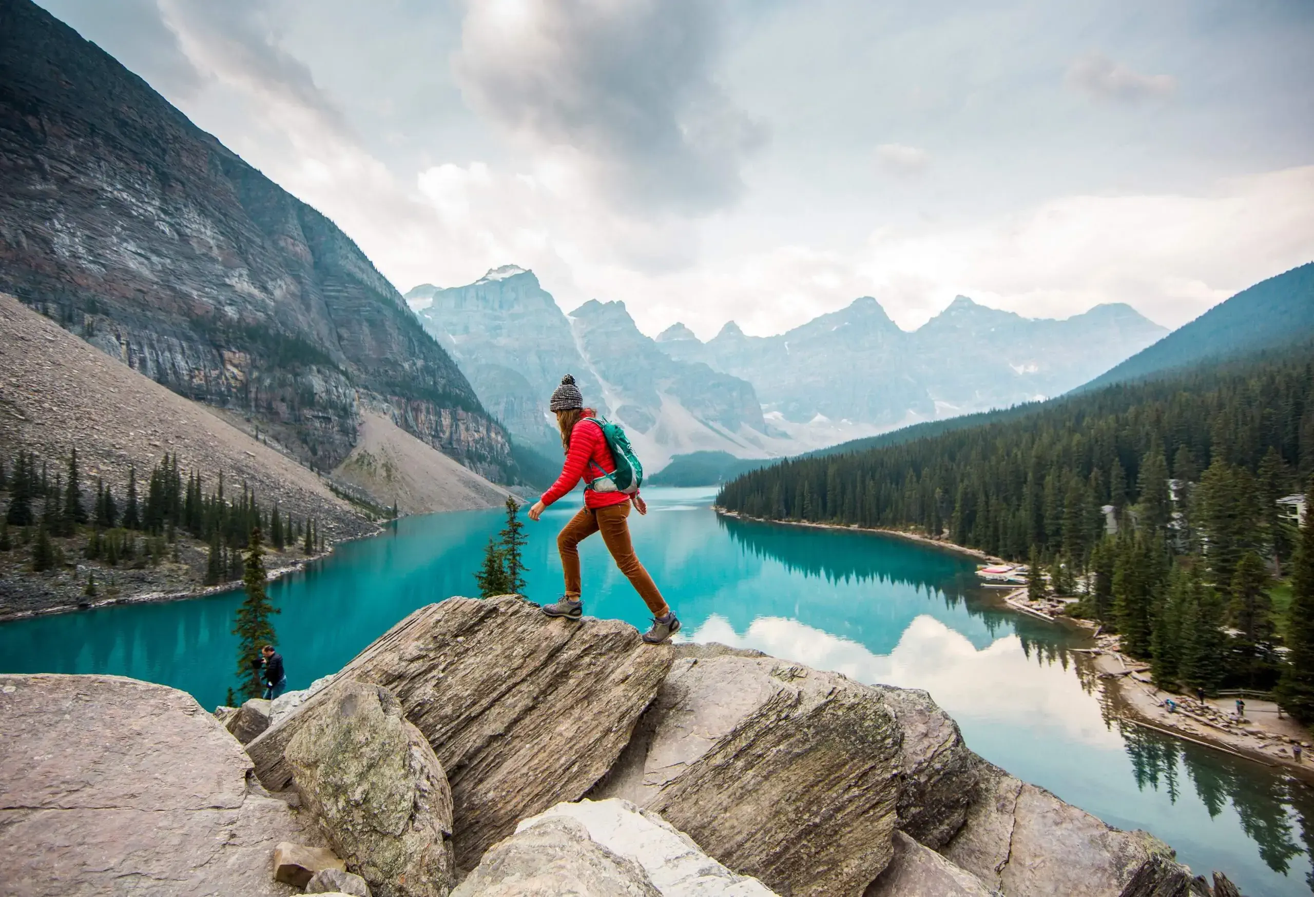 A hiker walks on a rocky hill's summit overlooking a calm lake bordered by a dense forest and vast mountain range.