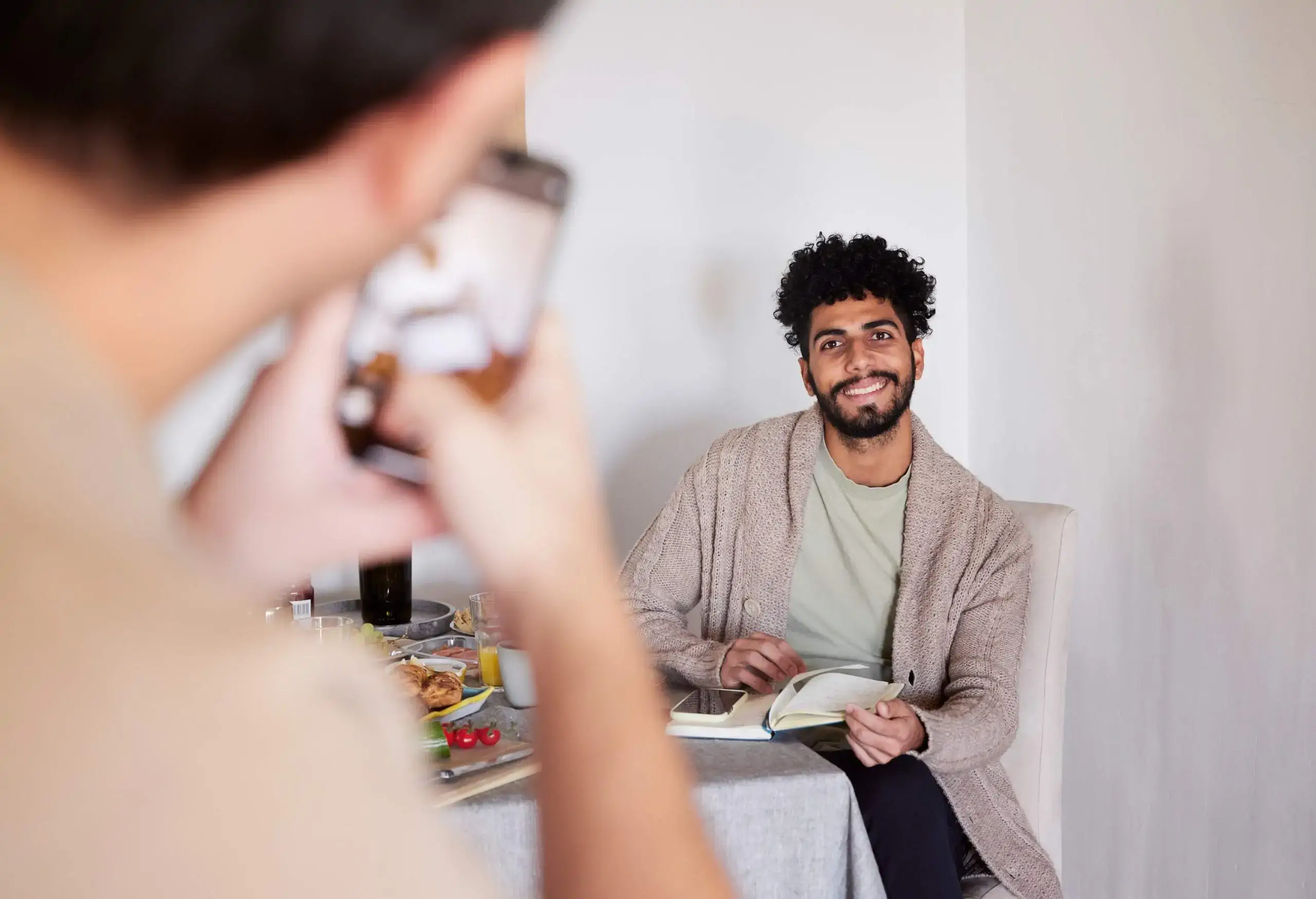 Smiling man sitting at table with open book, his partner taking picture with cell phone in foreground