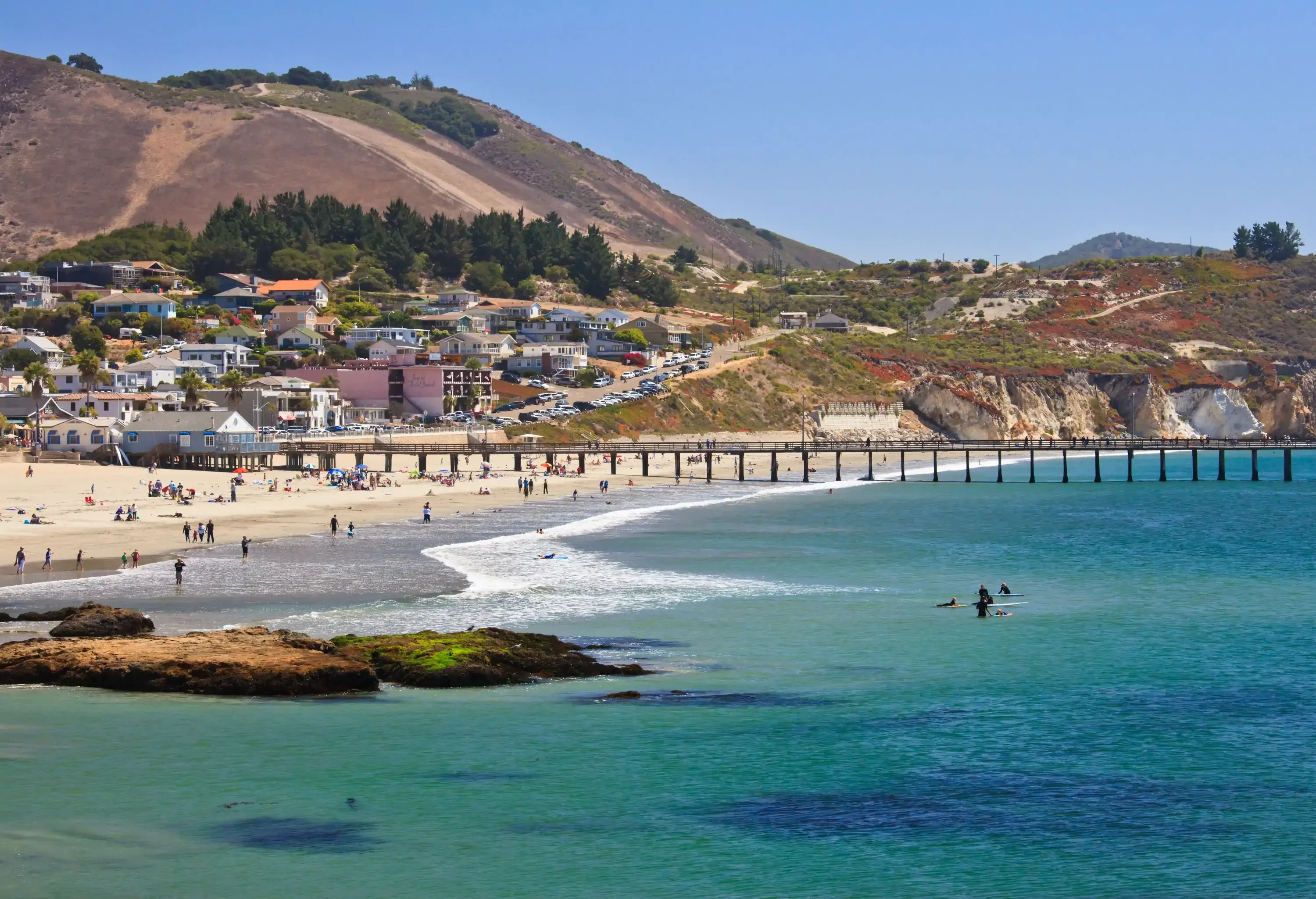 A quaint, spacious white sand beach with a long pier extending to the sea next to a seaside fishing village.