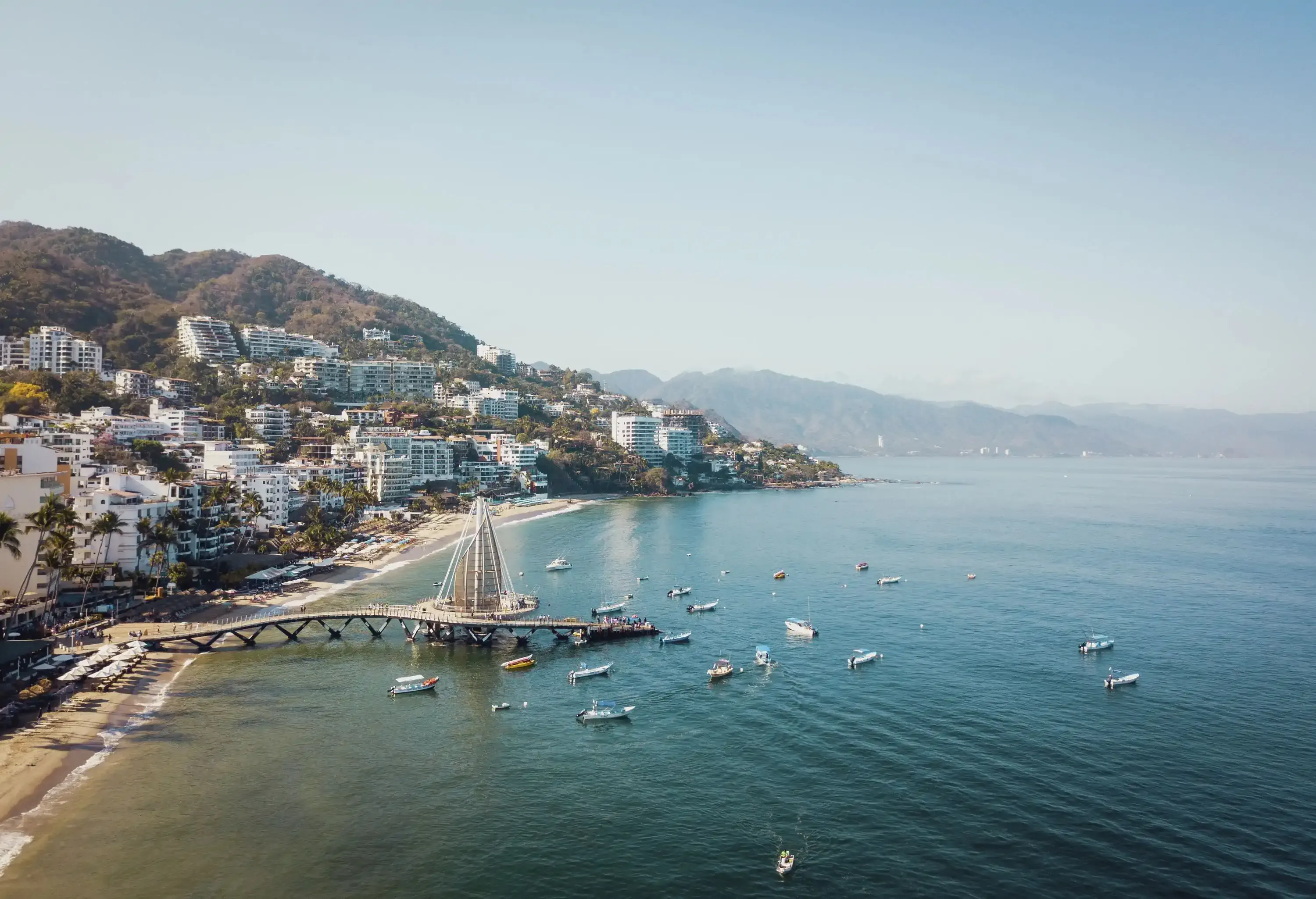 A picturesque coastal town boasts a bustling pier with vividly coloured boats, while buildings cascade down the hillside, embraced by verdant surroundings.