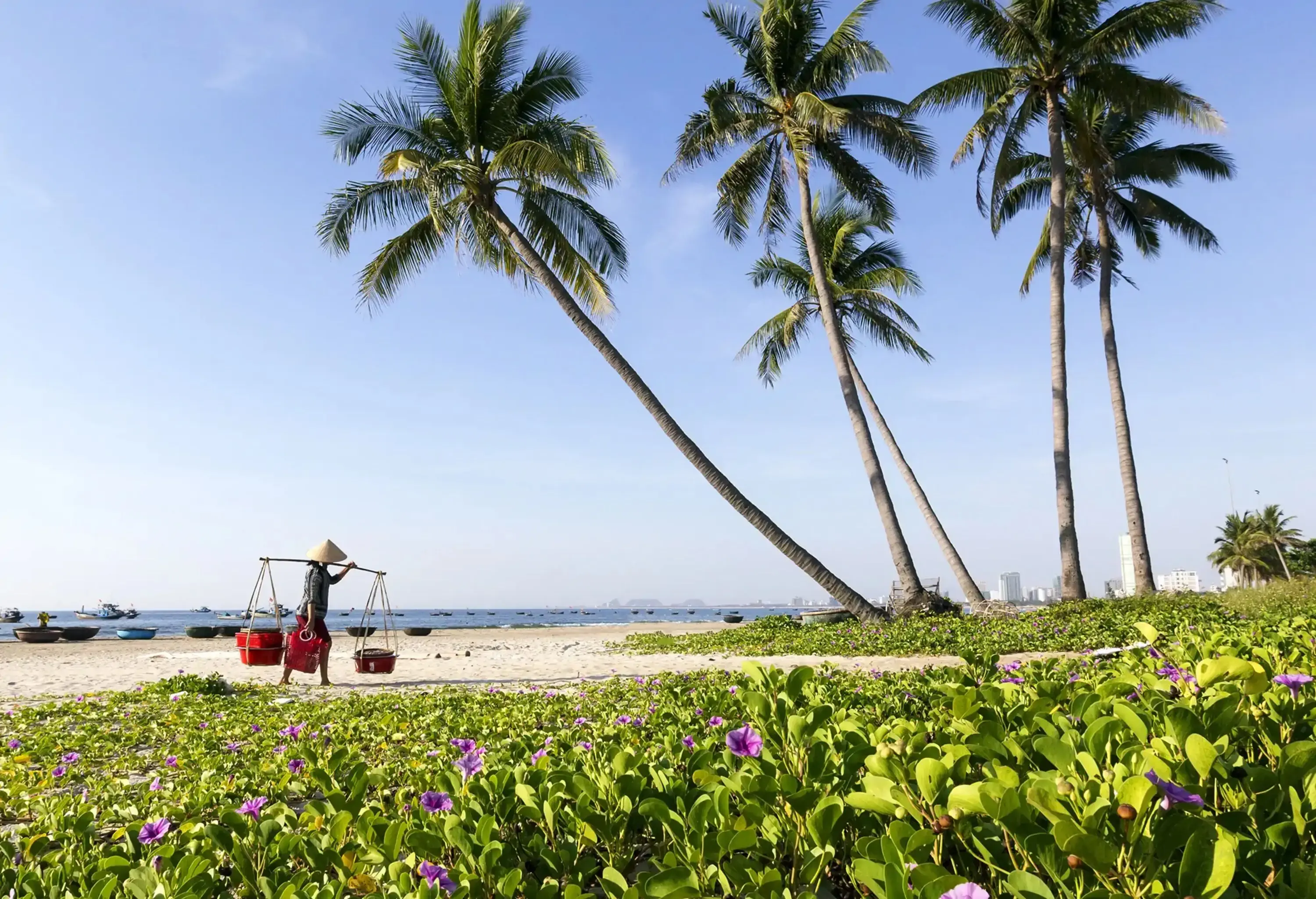 A man carrying baskets with a shoulder pole as he walks toward a beach covered in creeping vines next to a grove of palm trees.