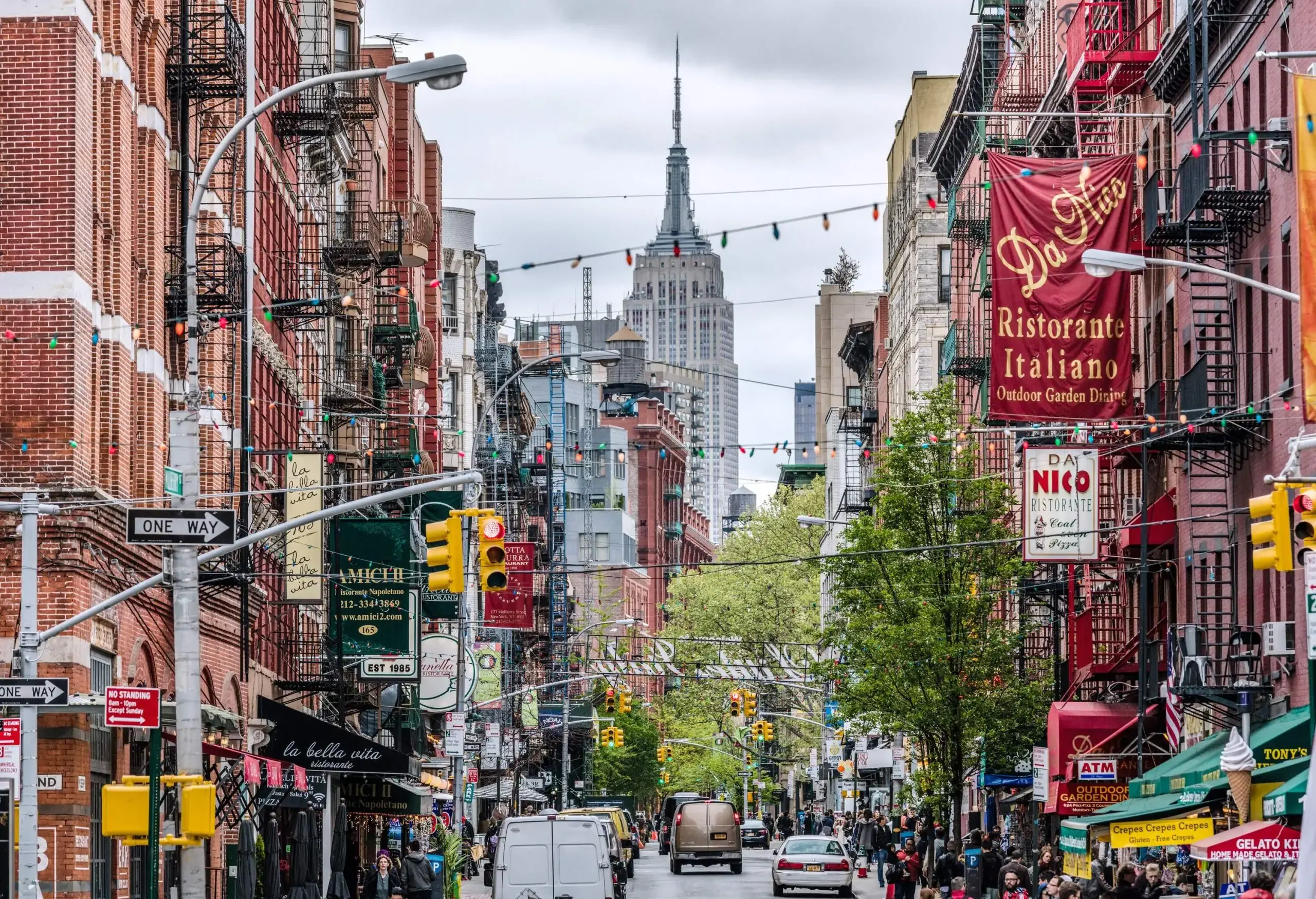 A view of the Empire State Building from a busy street in Little Italy, with buildings on both sides and cars driving past.