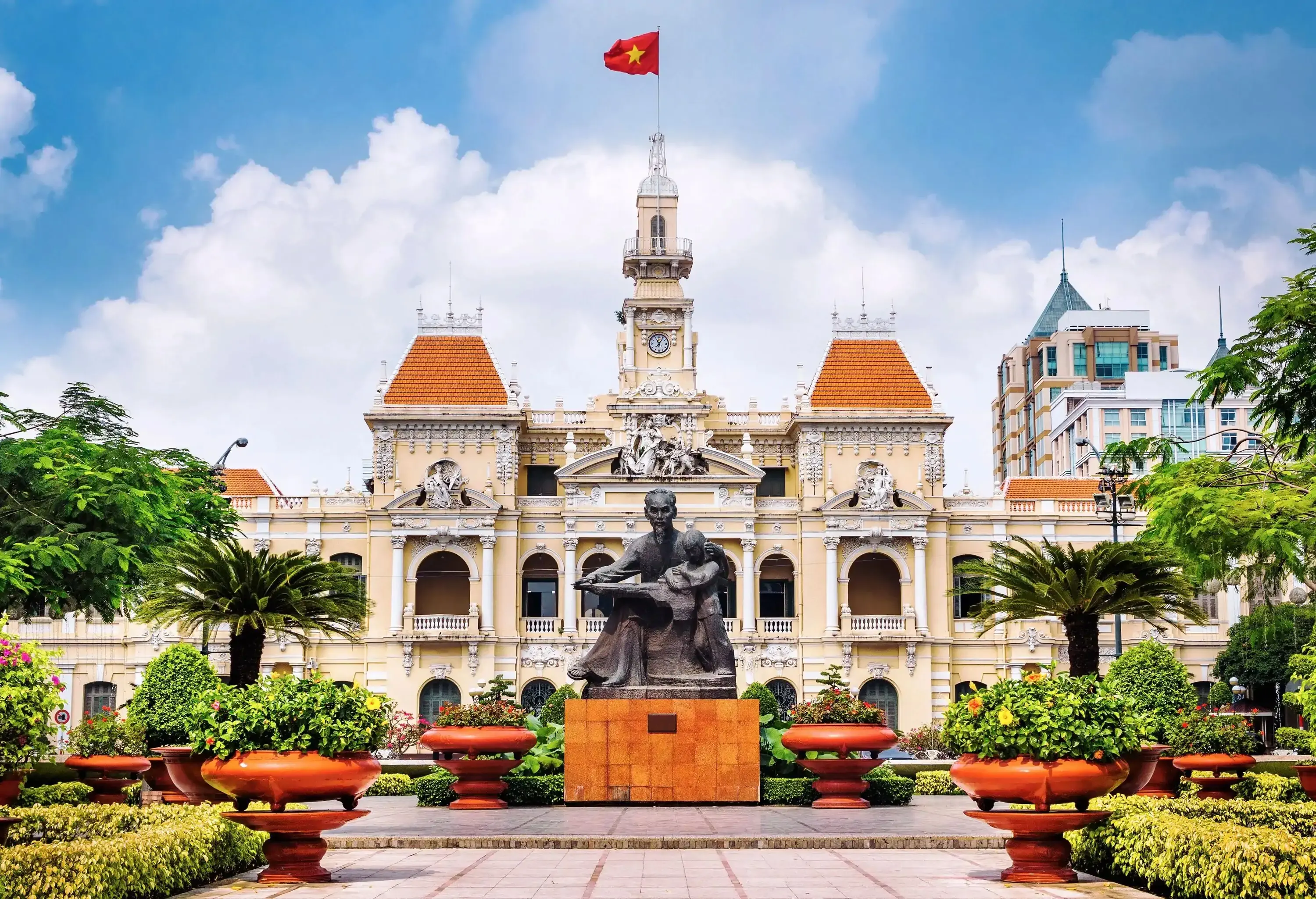 A statue in a curated garden with lush foliage in front of a palace with pillared balconies and a central clock tower.